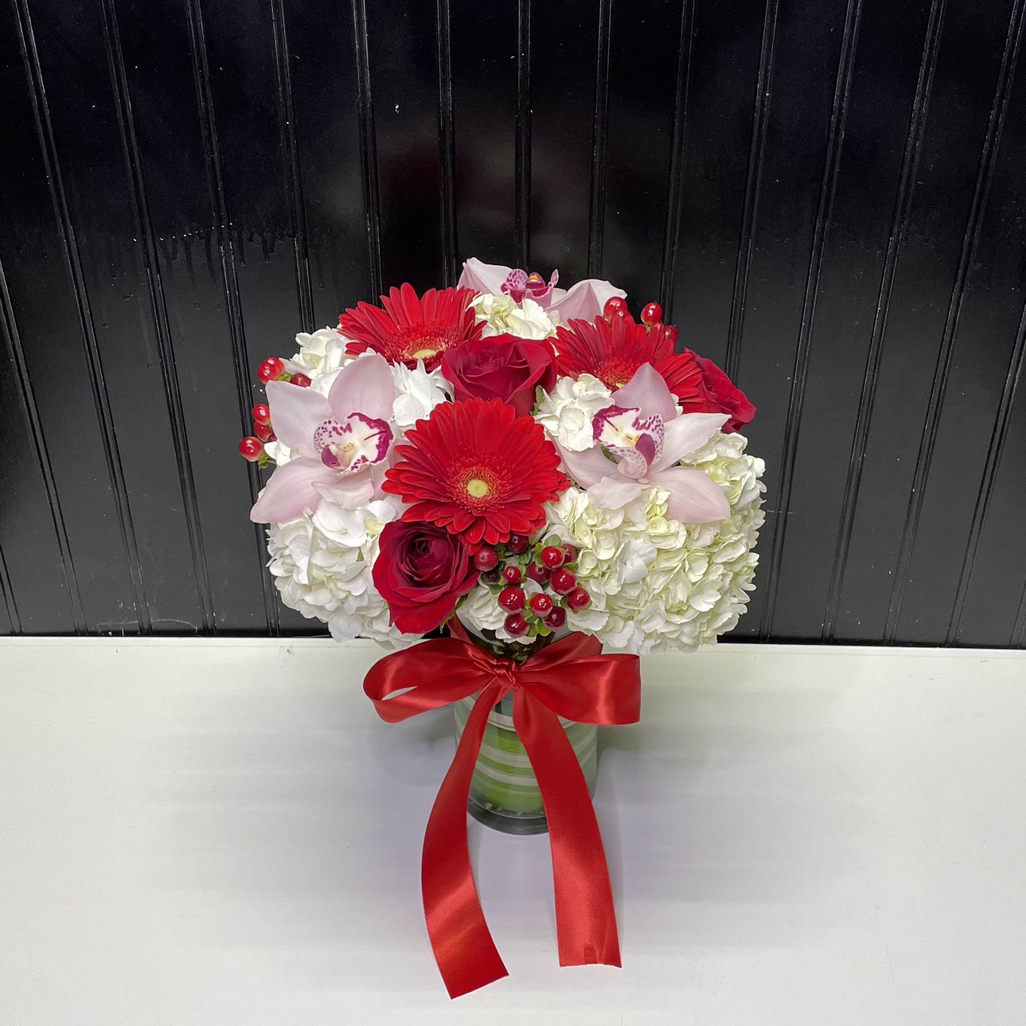 Round arrangement of red gerberas, roses, pink orchids and white hydrangeas in a glass vase with red bow.