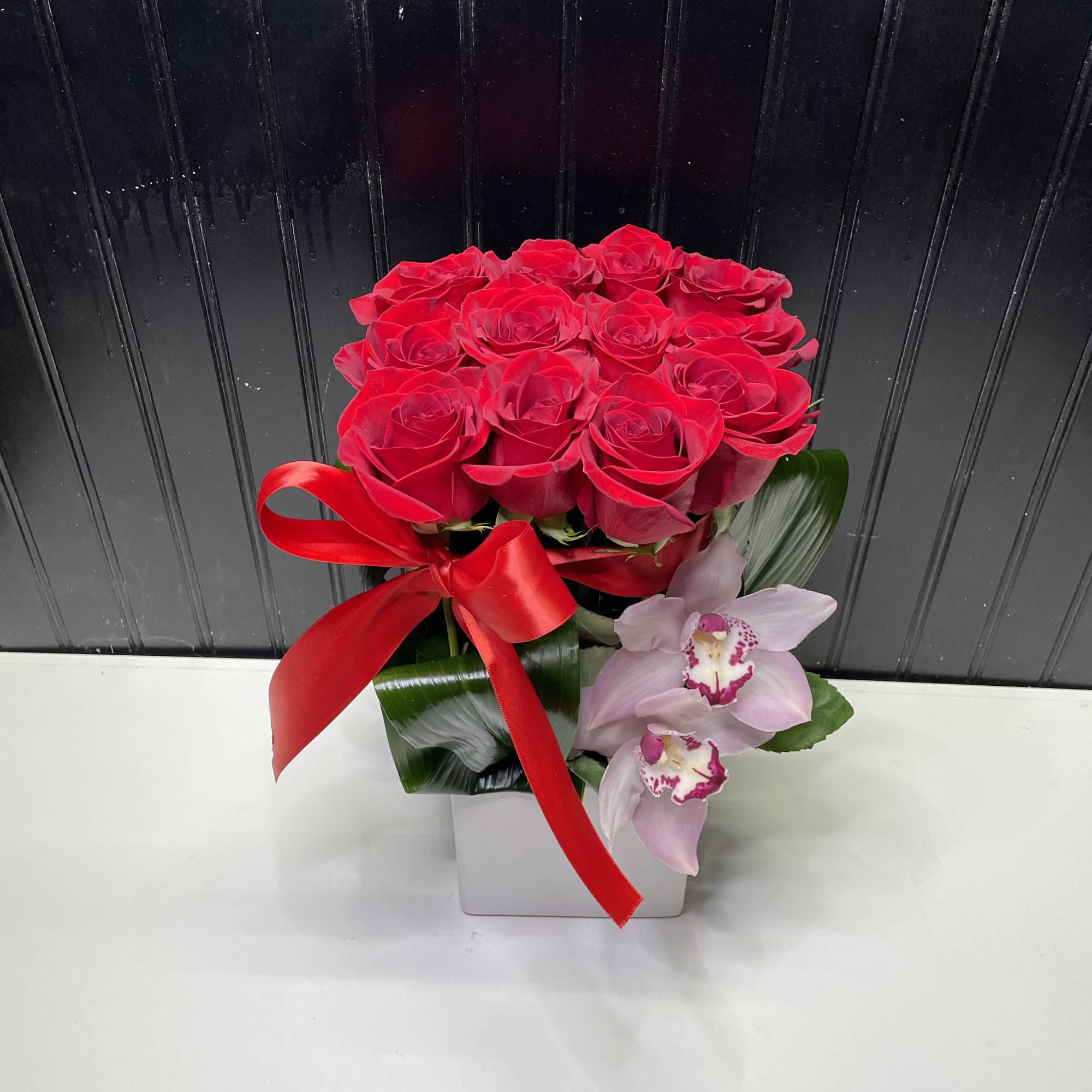 Compact arrangement of red roses and pink orchids in a white square vase with a red ribbon bow.