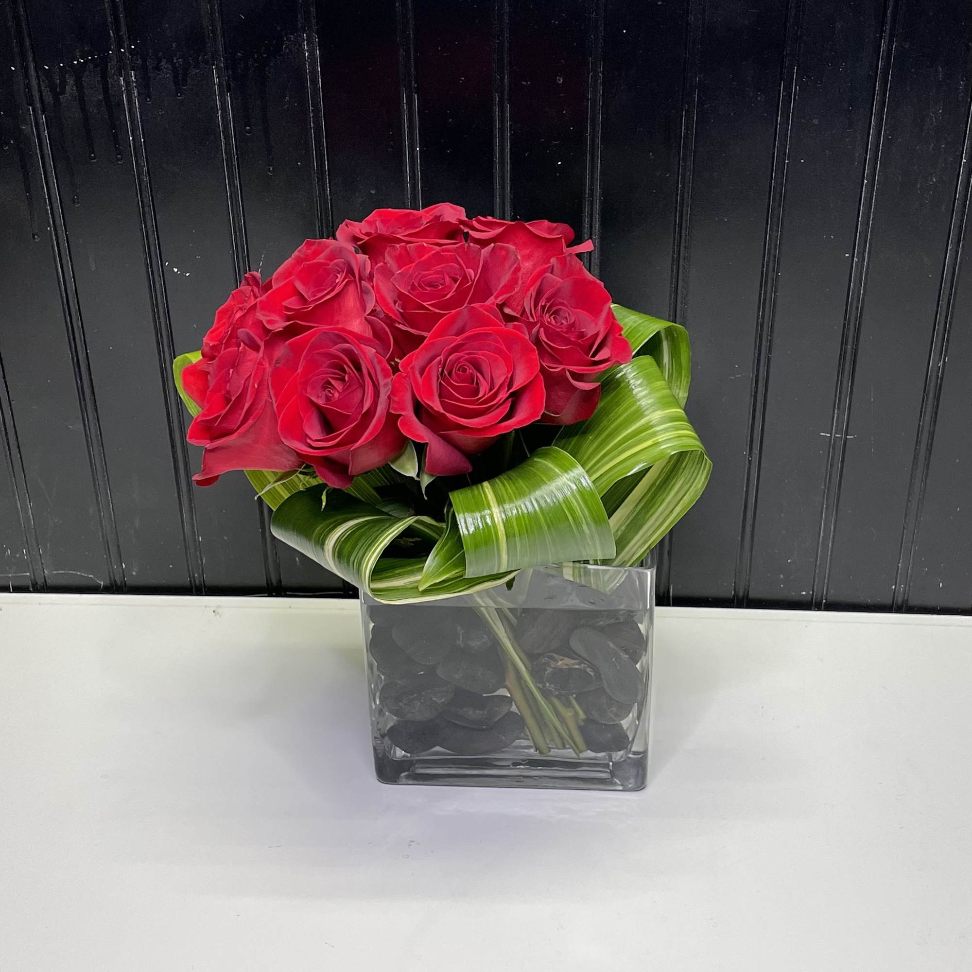 Compact arrangement of red roses with looped green foliage in a square glass vase with black stones