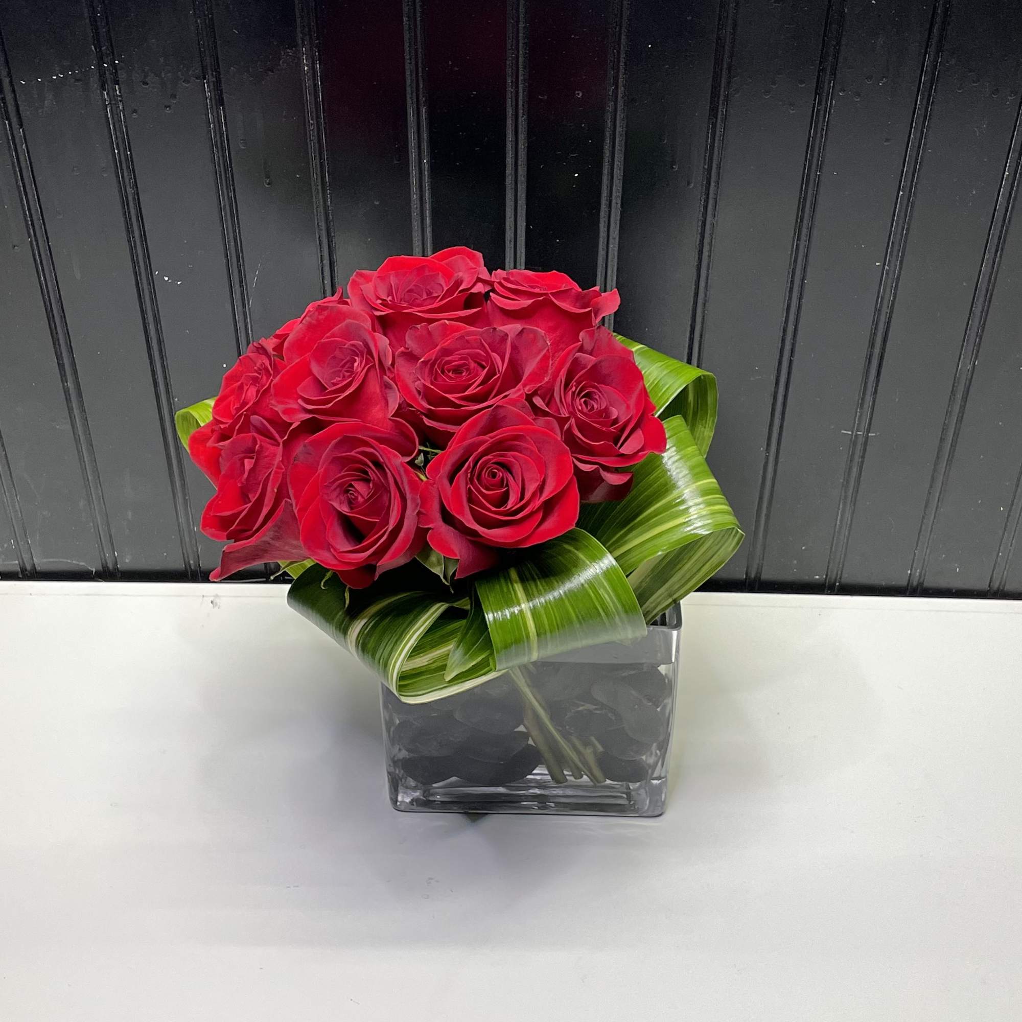 Compact arrangement of red roses in a square glass vase with a collar of folded green leaves.