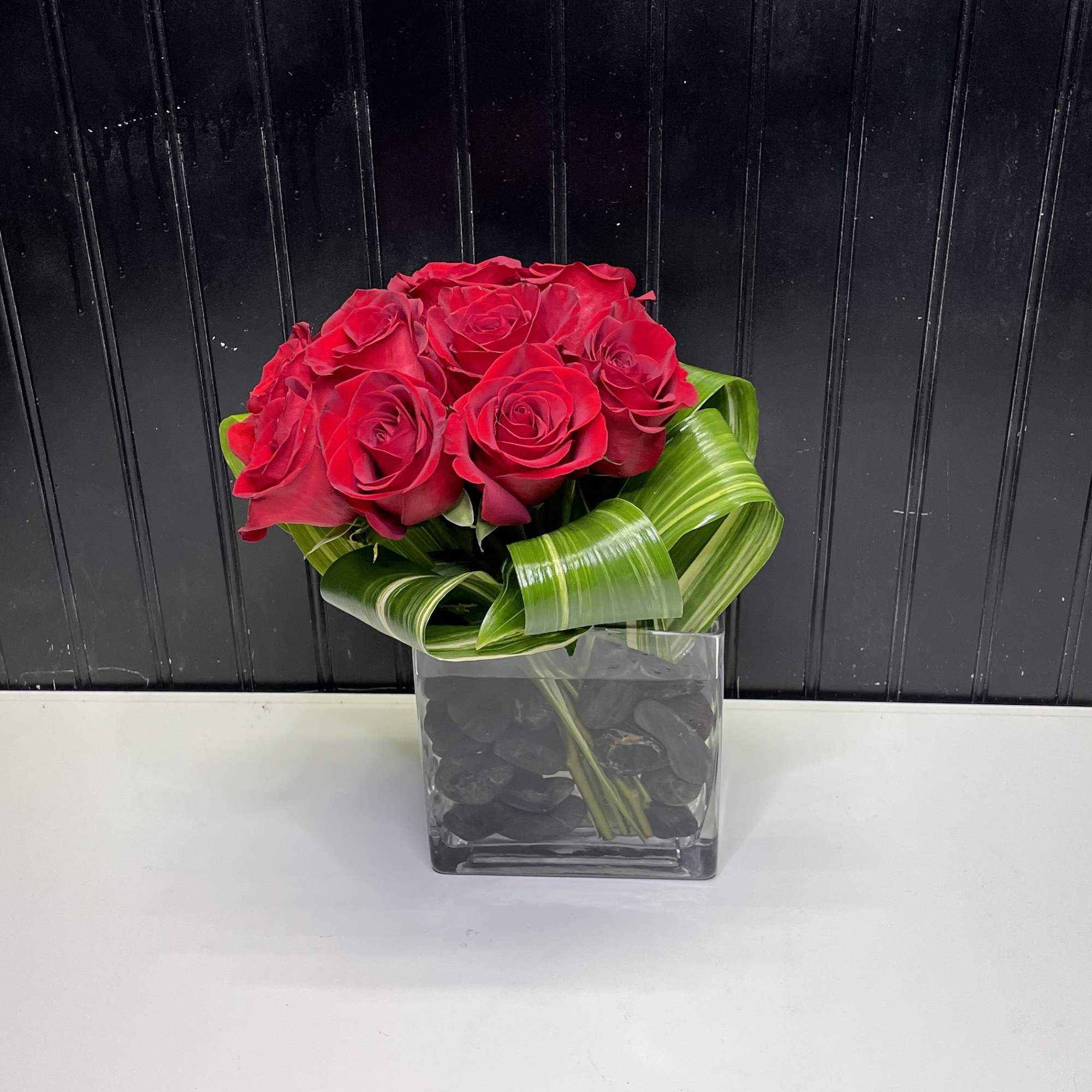 Compact arrangement of red roses in a square glass vase with folded green leaf loops.