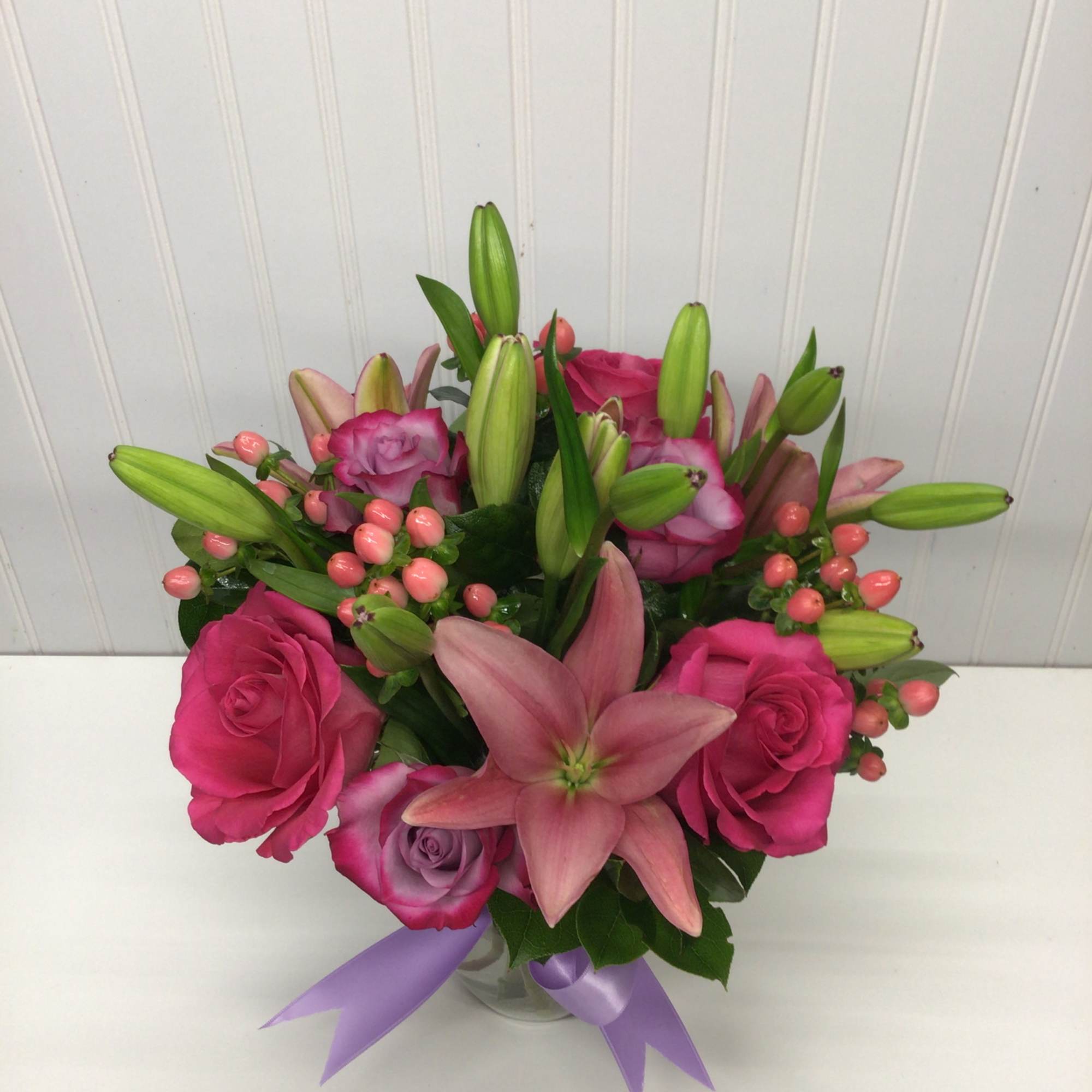 Pink lilies and roses arranged in a clear vase with a lavender ribbon bow