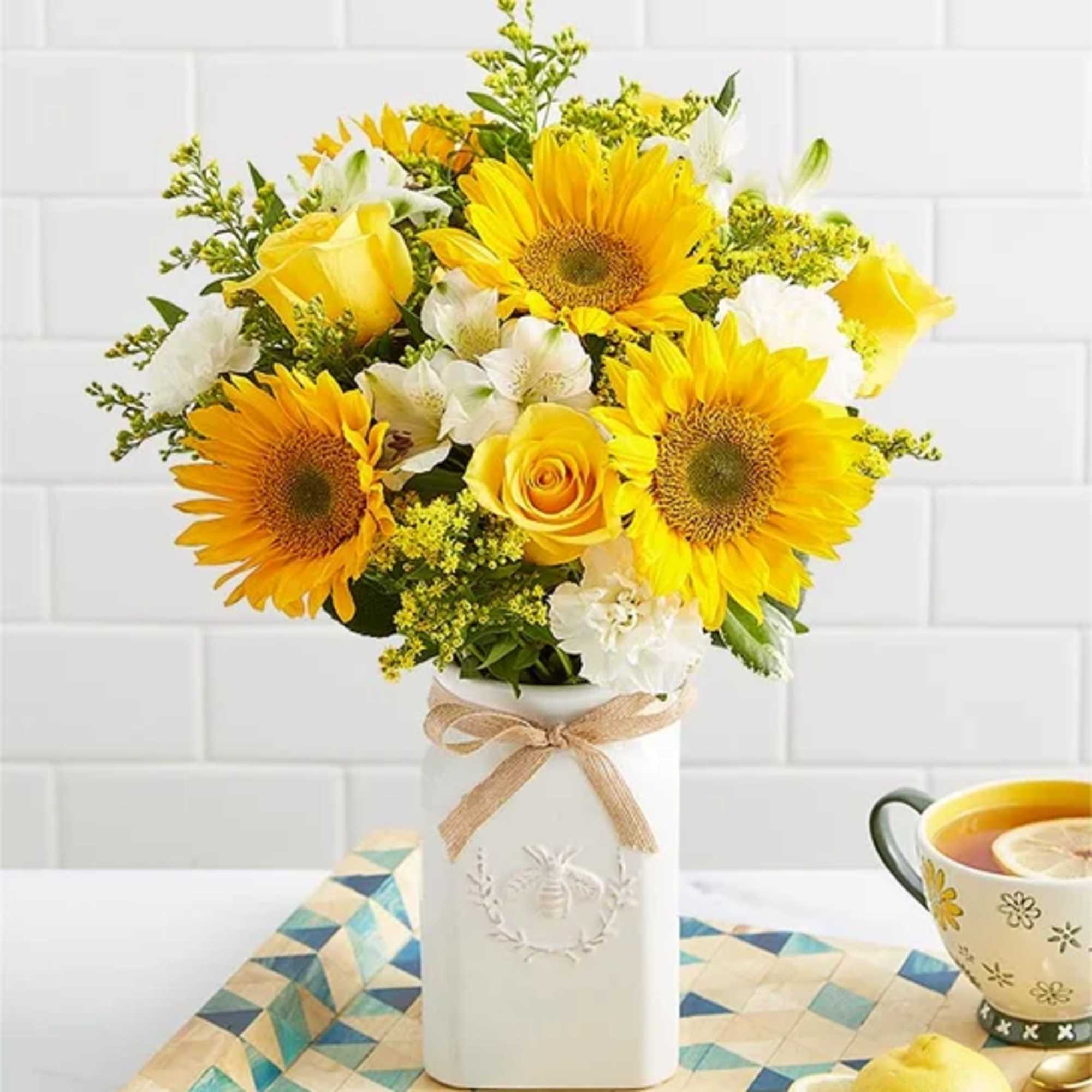 Tall white vase of yellow sunflowers, roses, and white blooms on a kitchen table