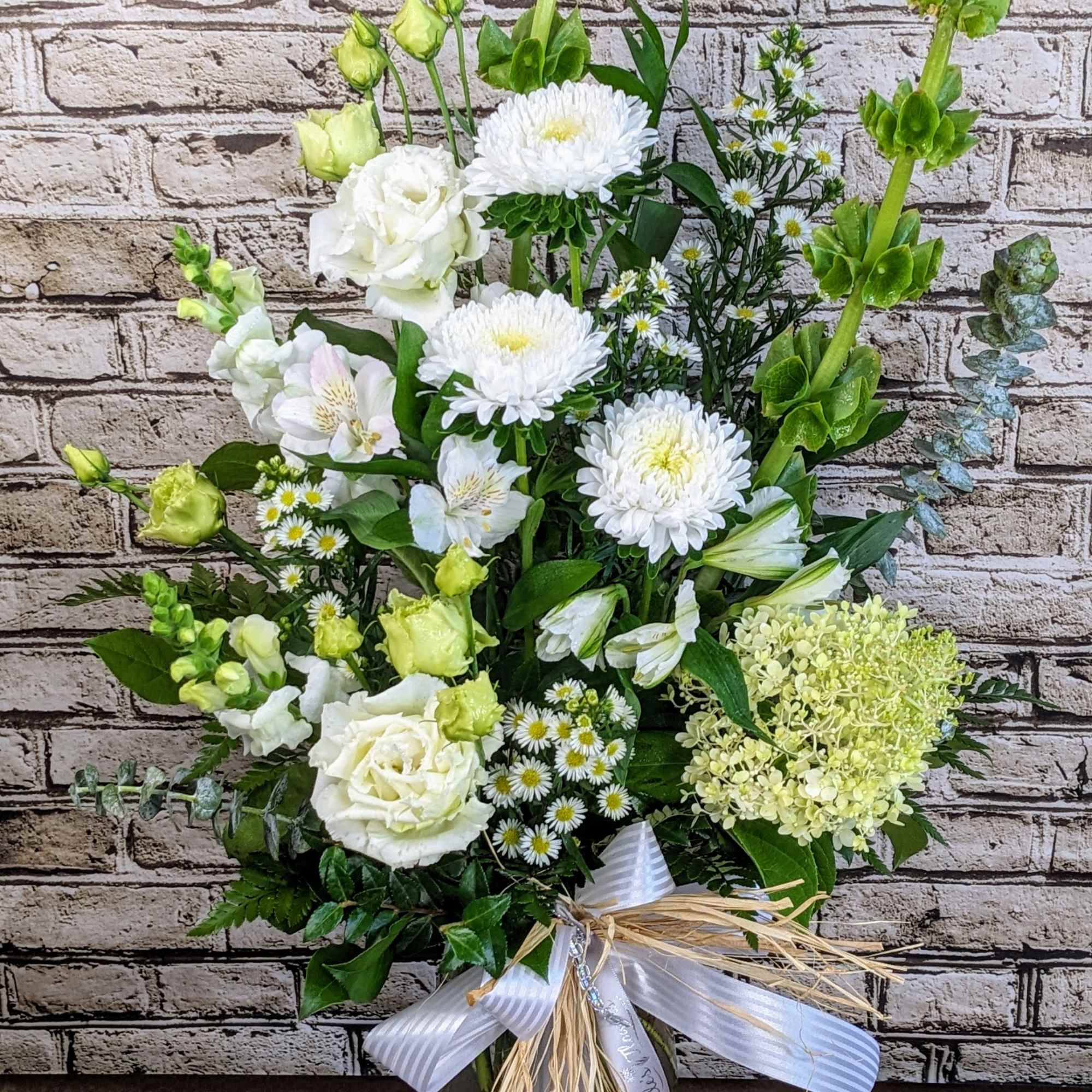 Tall white and green arrangement with roses, mums, and bells of Ireland in a glass vase with ribbon.