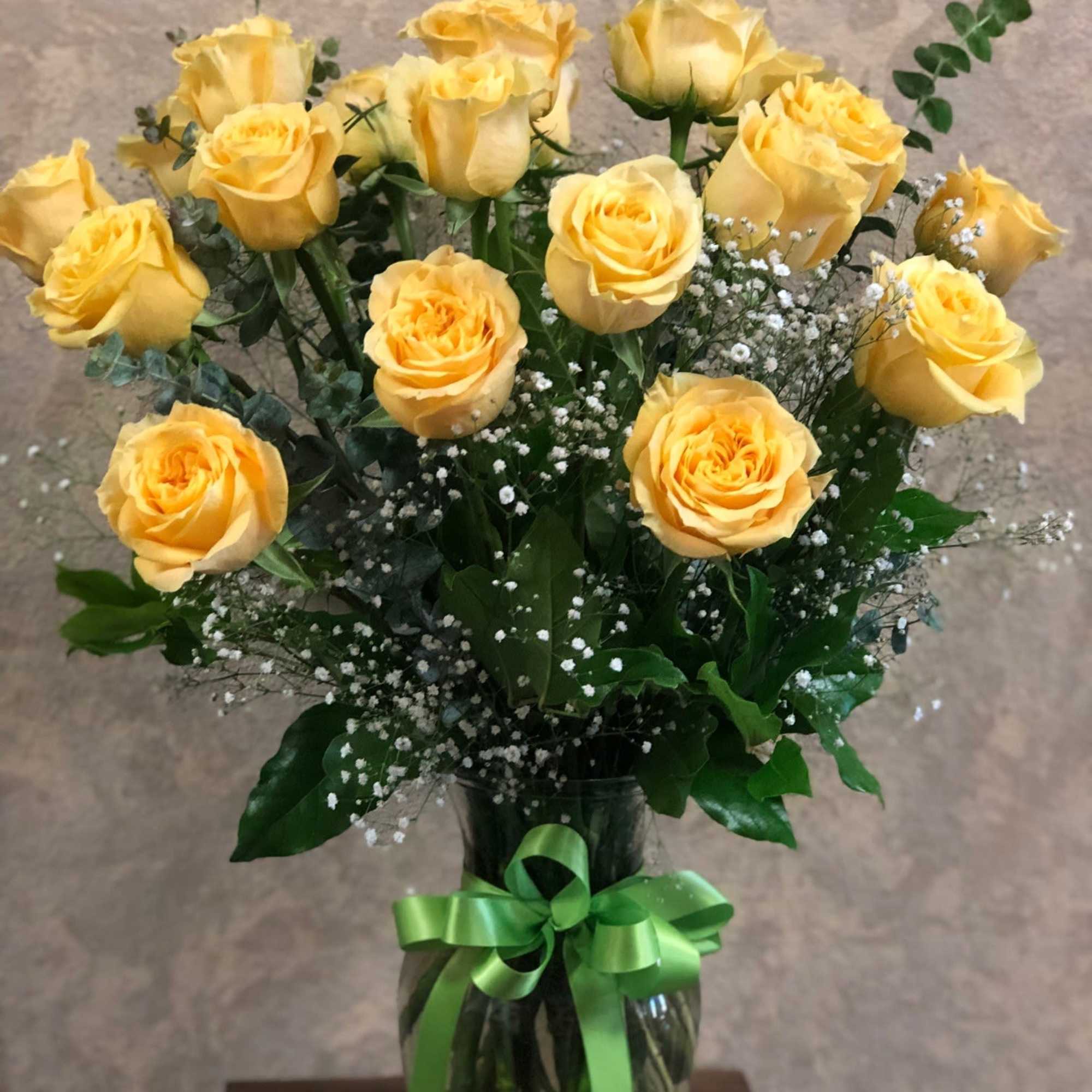 Arrangement of yellow roses with white filler flowers in a clear glass vase tied with a green ribbon.