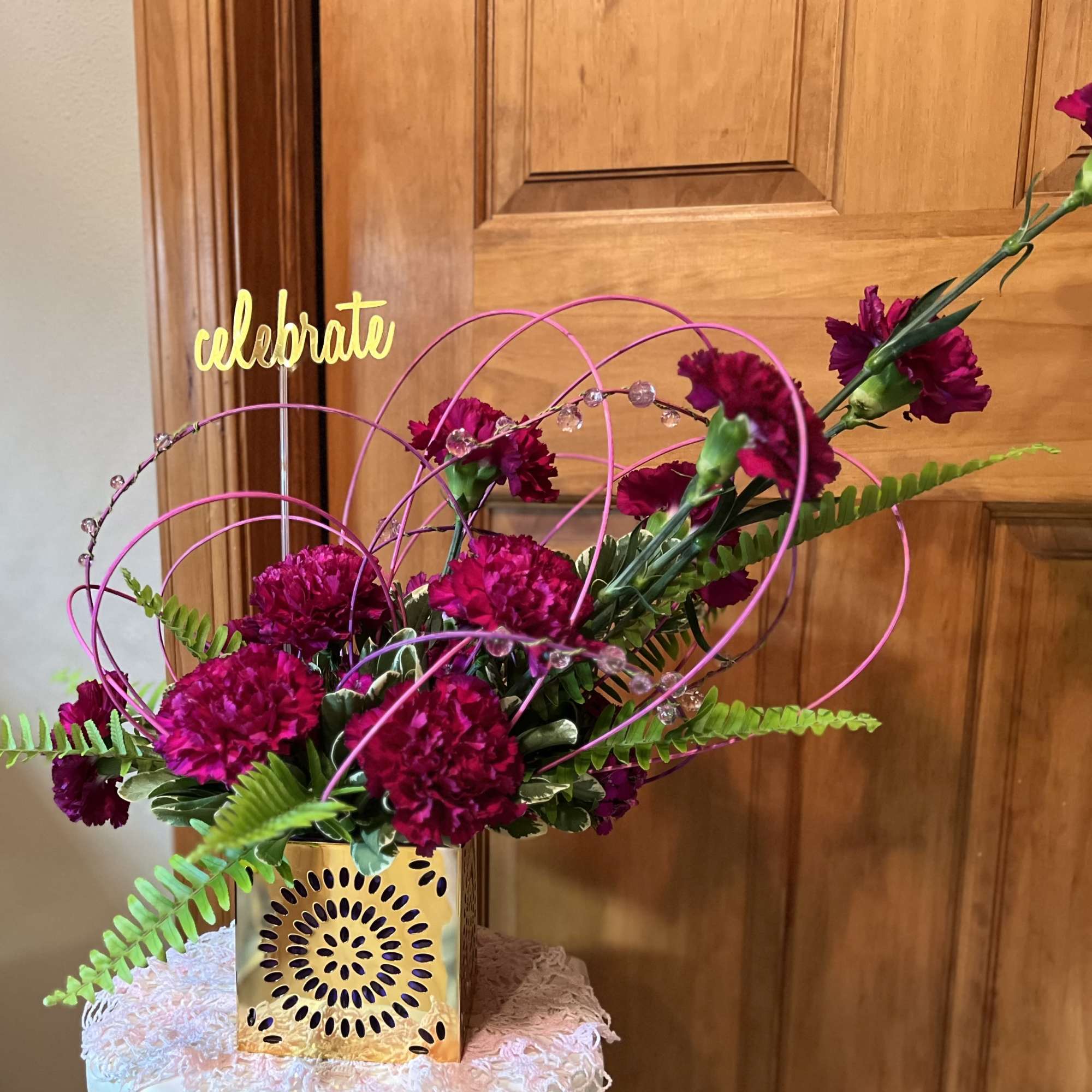 Magenta carnations in a decorative gold vase with pink wire loops and a "celebrate" sign