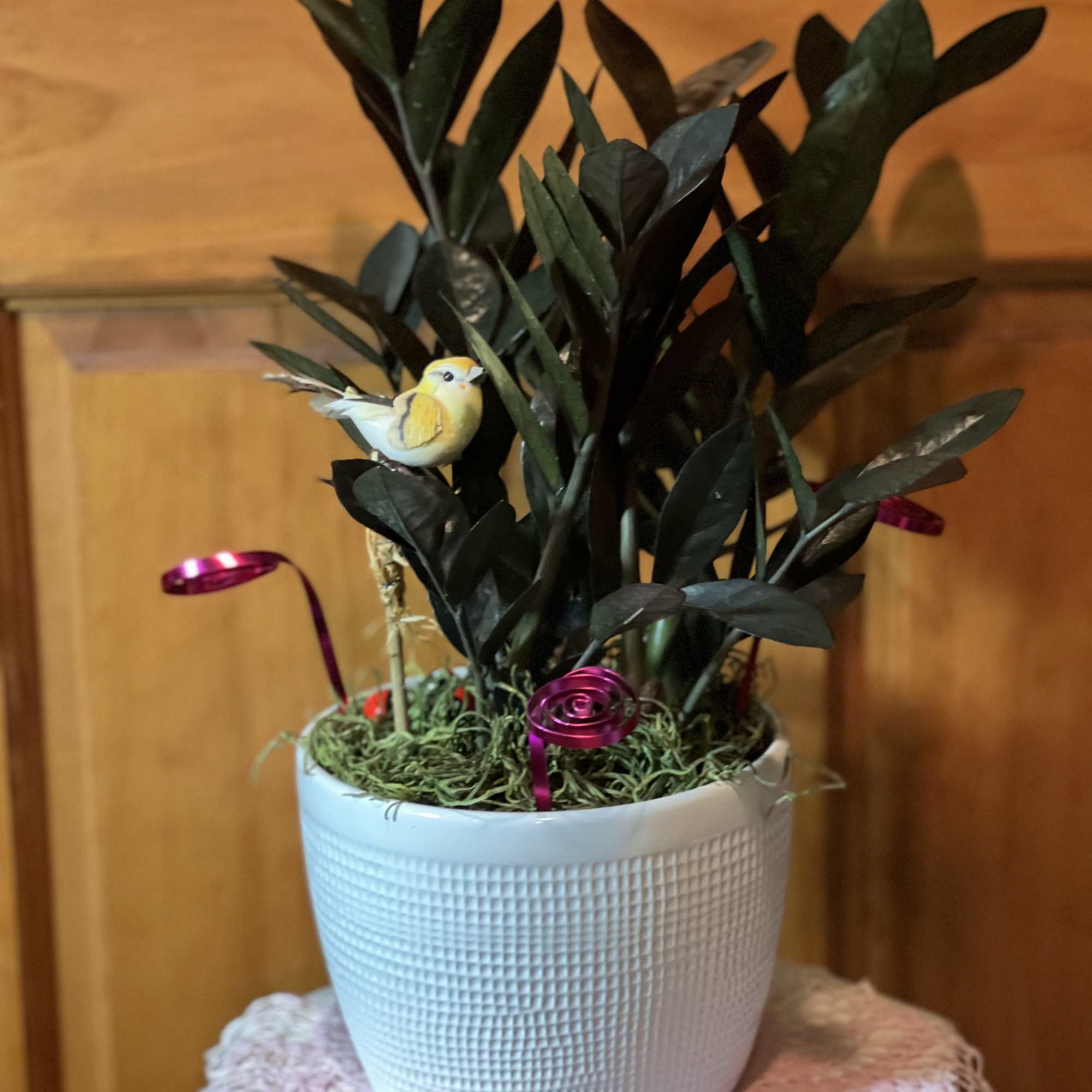Potted plant with dark leaves, a small bird ornament, and pink ribbon accents