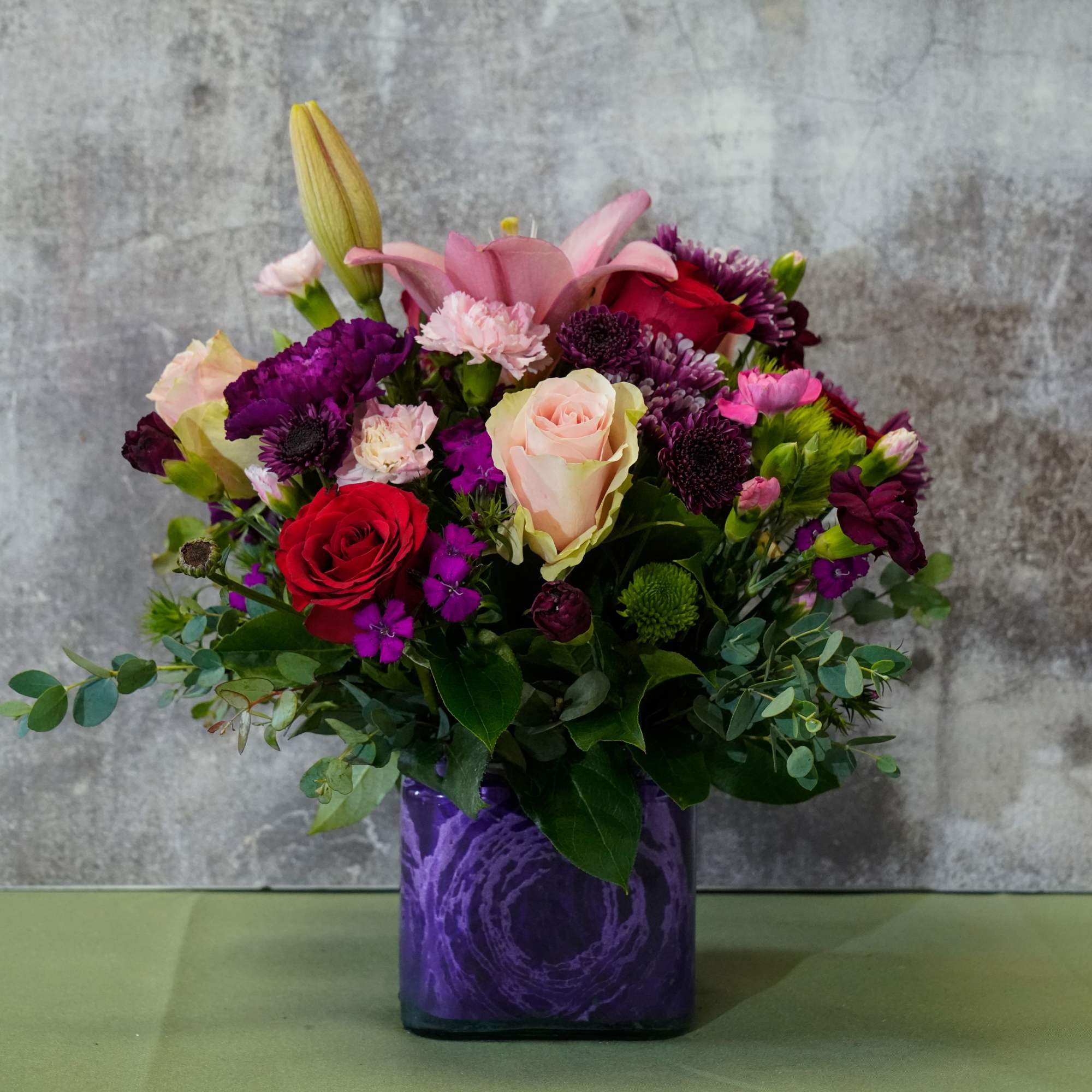 Mixed bouquet of roses, lilies, and purple flowers in a purple vase