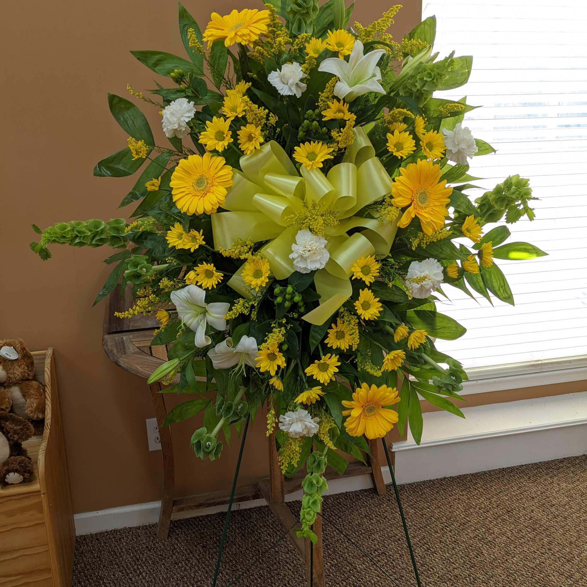 Standing floral spray with yellow daisies, white lilies, and a green ribbon bow