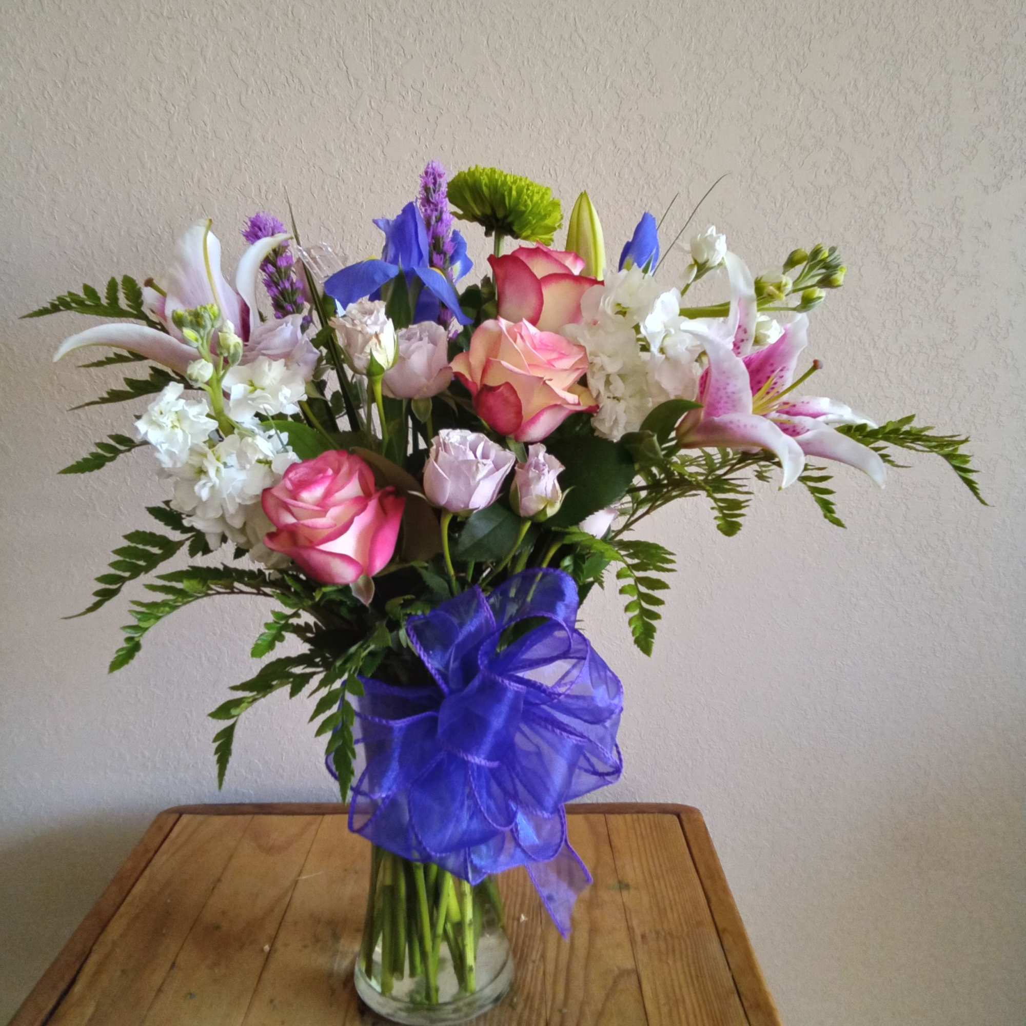 Mixed bouquet of roses, lilies, and blue flowers in a glass vase with a purple bow