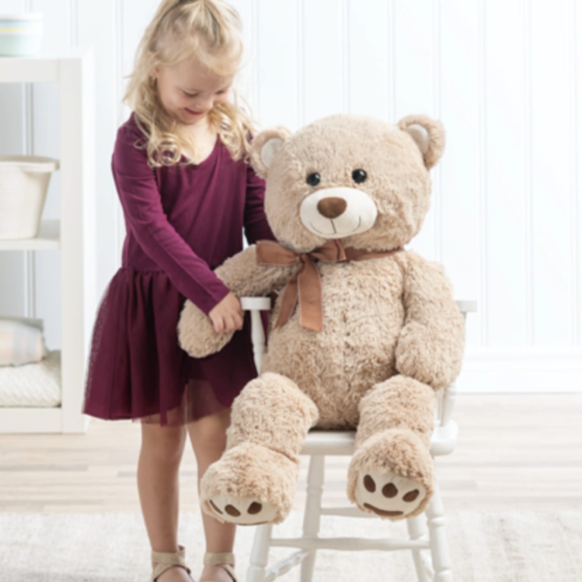 Large tan teddy bear with bow sitting on white chair beside a young girl in a burgundy dress.