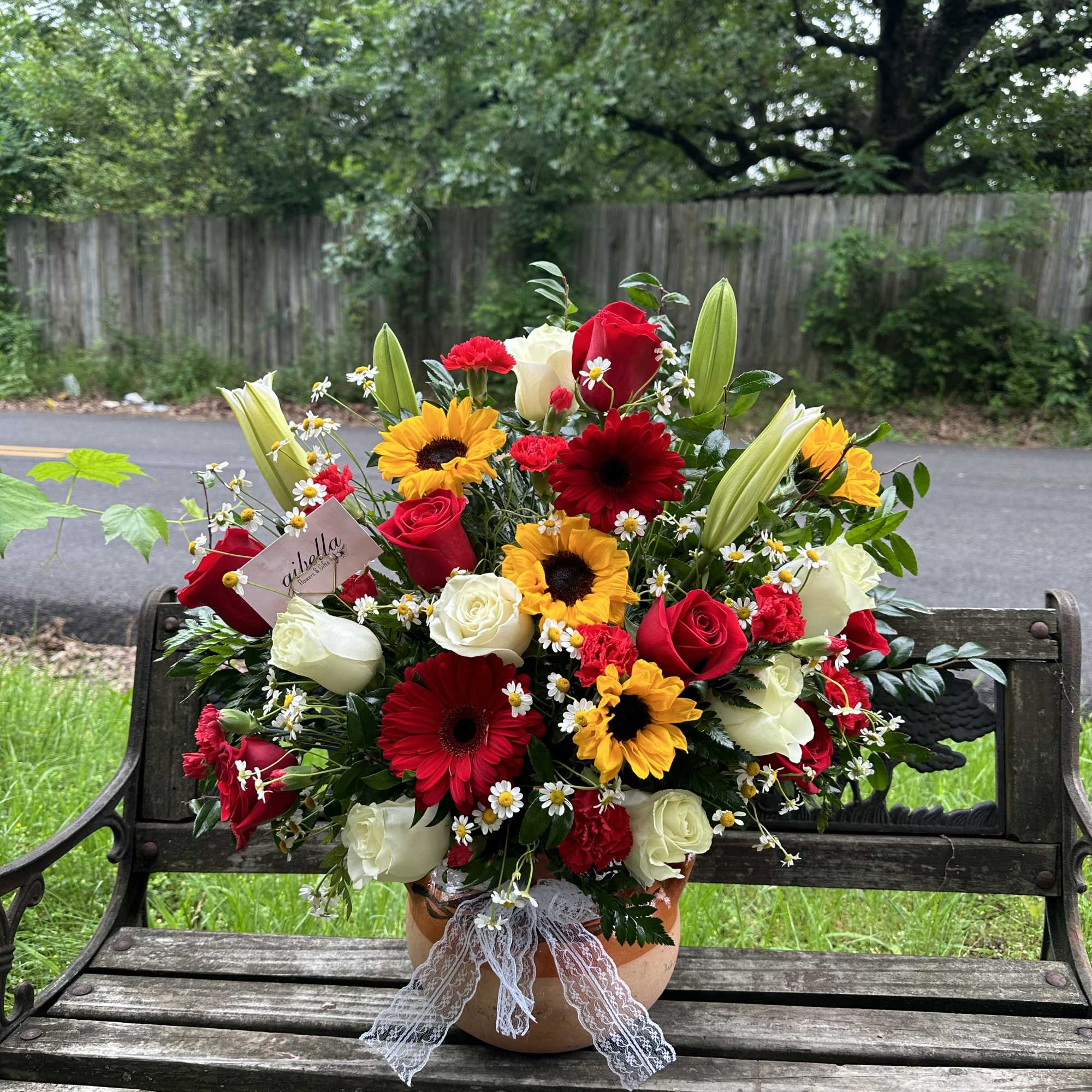 Large mixed arrangement of red and white roses, sunflowers, gerbera daisies, and lilies in a pot with a lace bow.
