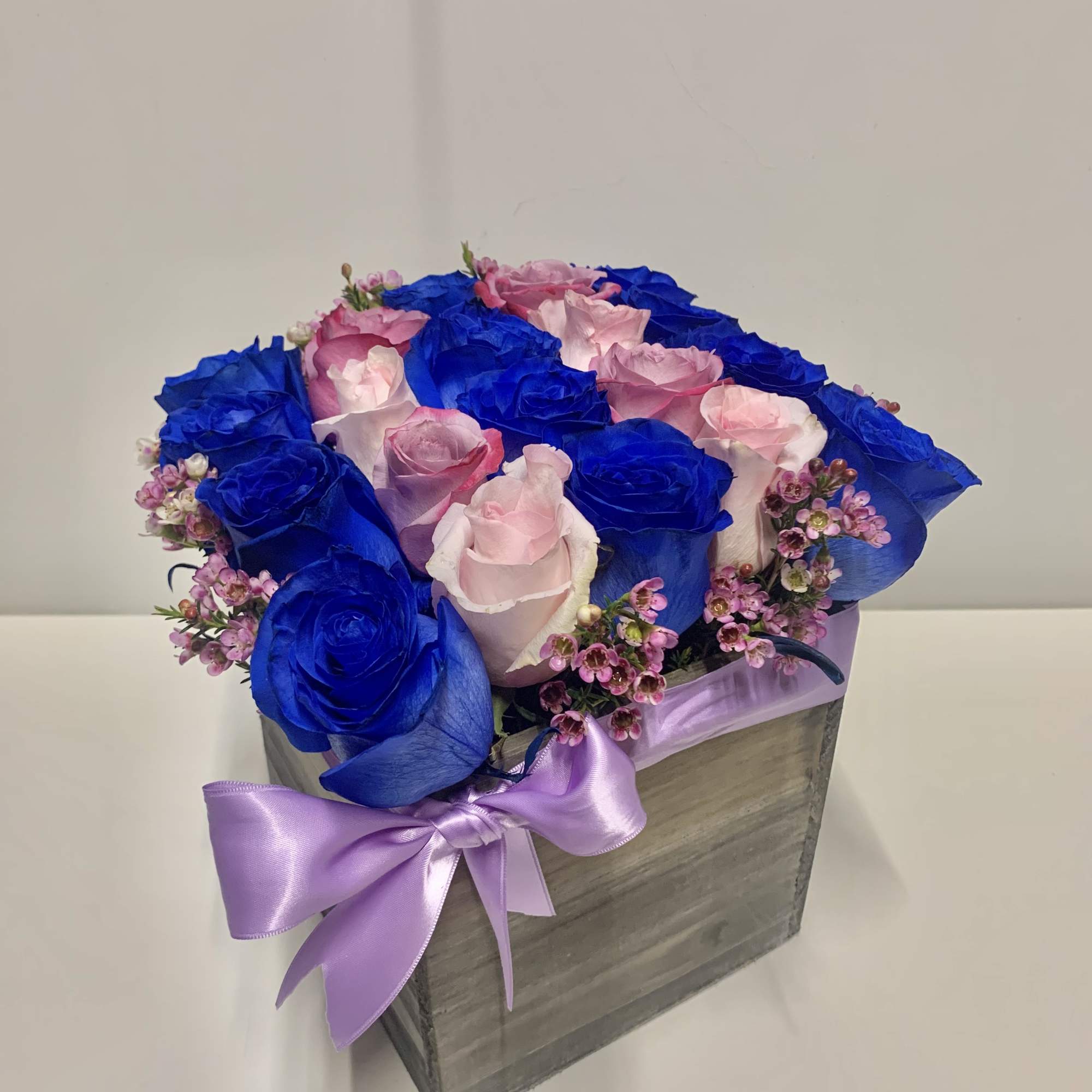 Square wooden box of bright blue and light pink roses with tiny pink blooms and a lavender bow.