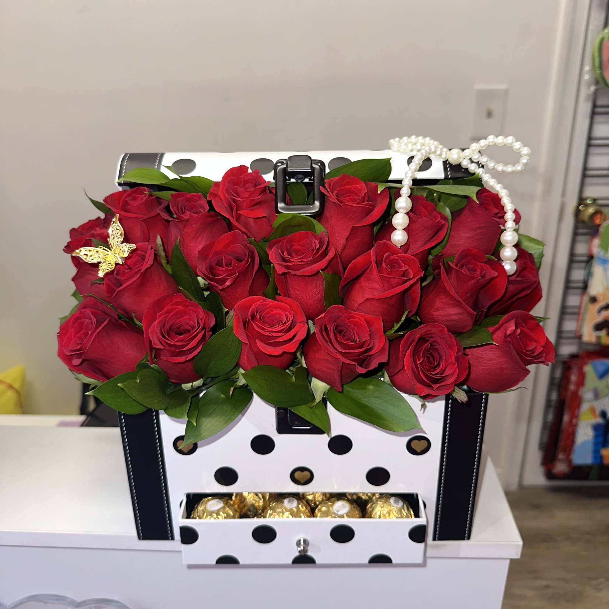 Red roses arranged in a polka-dot gift box with a pearl handle and chocolates below.