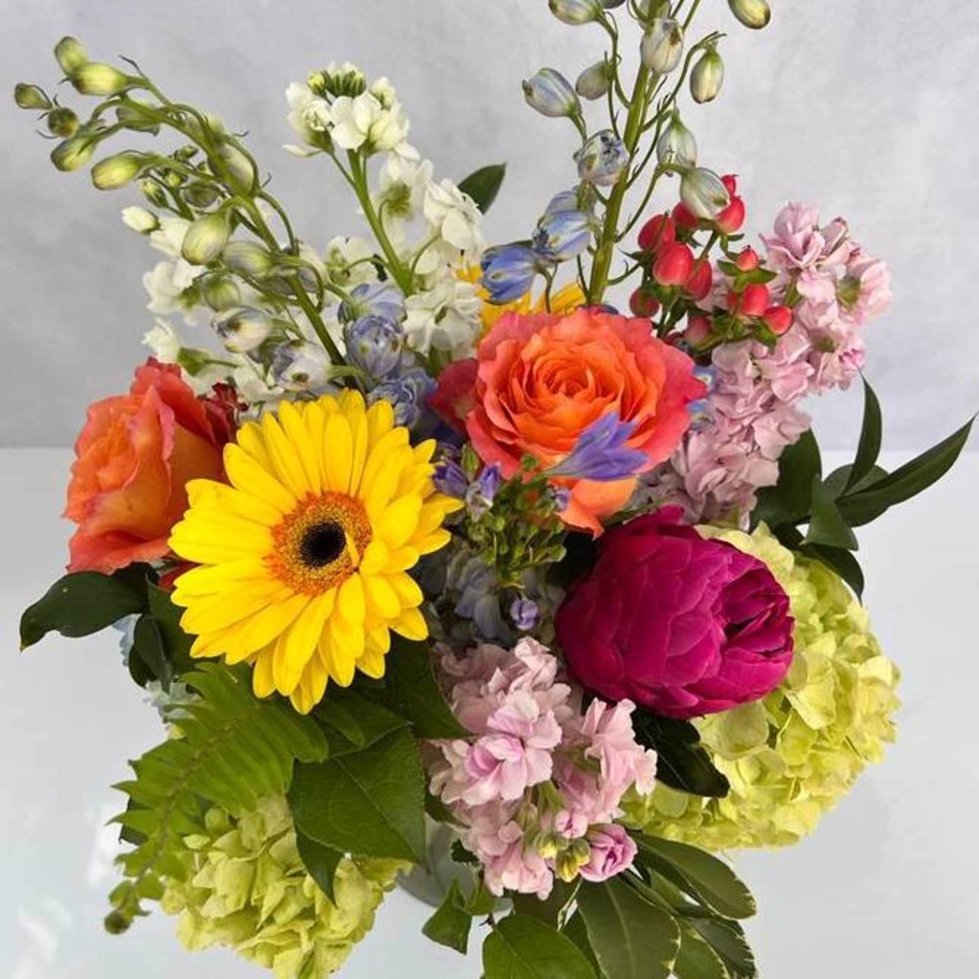 Vibrant mixed arrangement with yellow gerbera daisy, orange roses, pink peony, and green hydrangeas in a vase