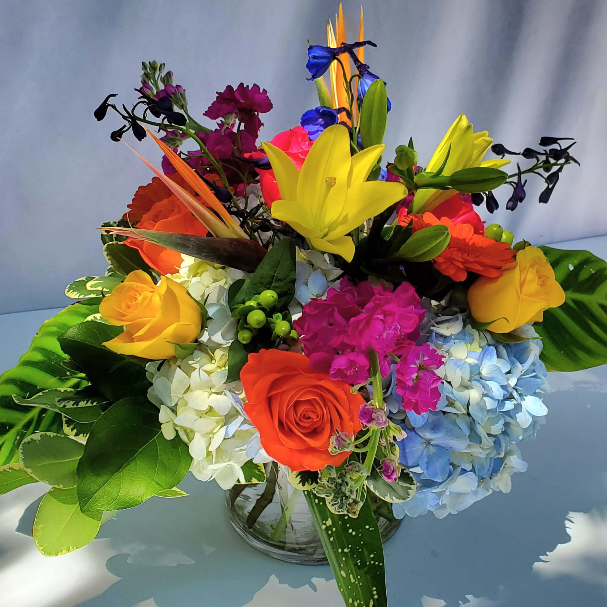 Colorful low arrangement with yellow lilies, orange and yellow roses, and blue hydrangeas in a clear vase