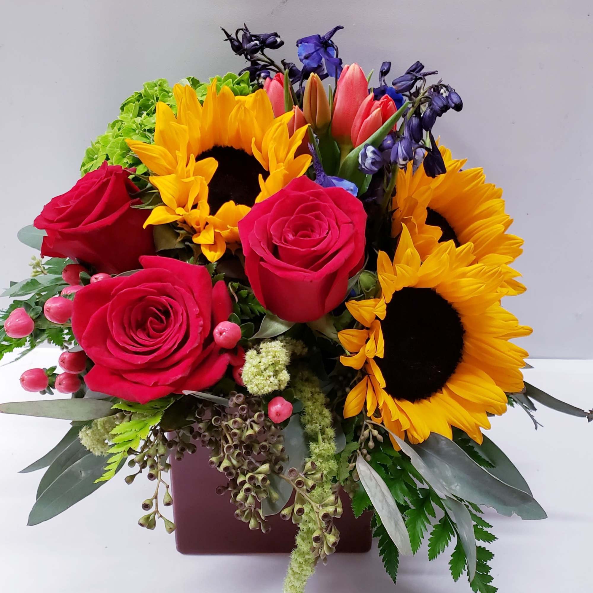 Bouquet of red roses, sunflowers, and purple flowers in a square vase
