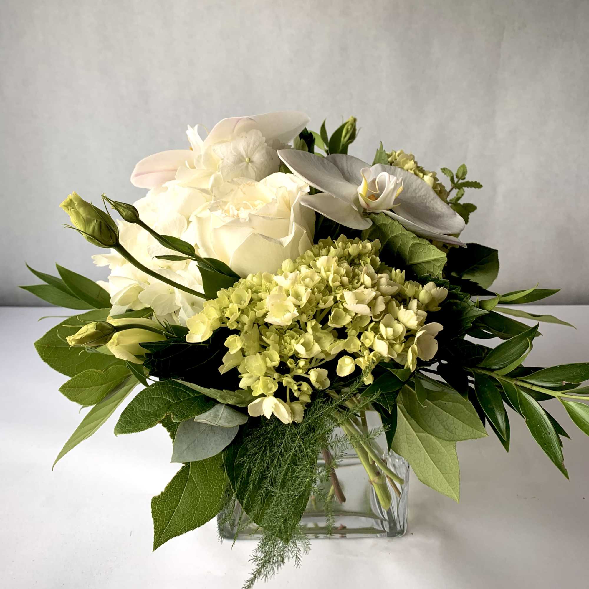 Low arrangement of white roses, orchids, and pale green hydrangeas in a clear glass cube vase.