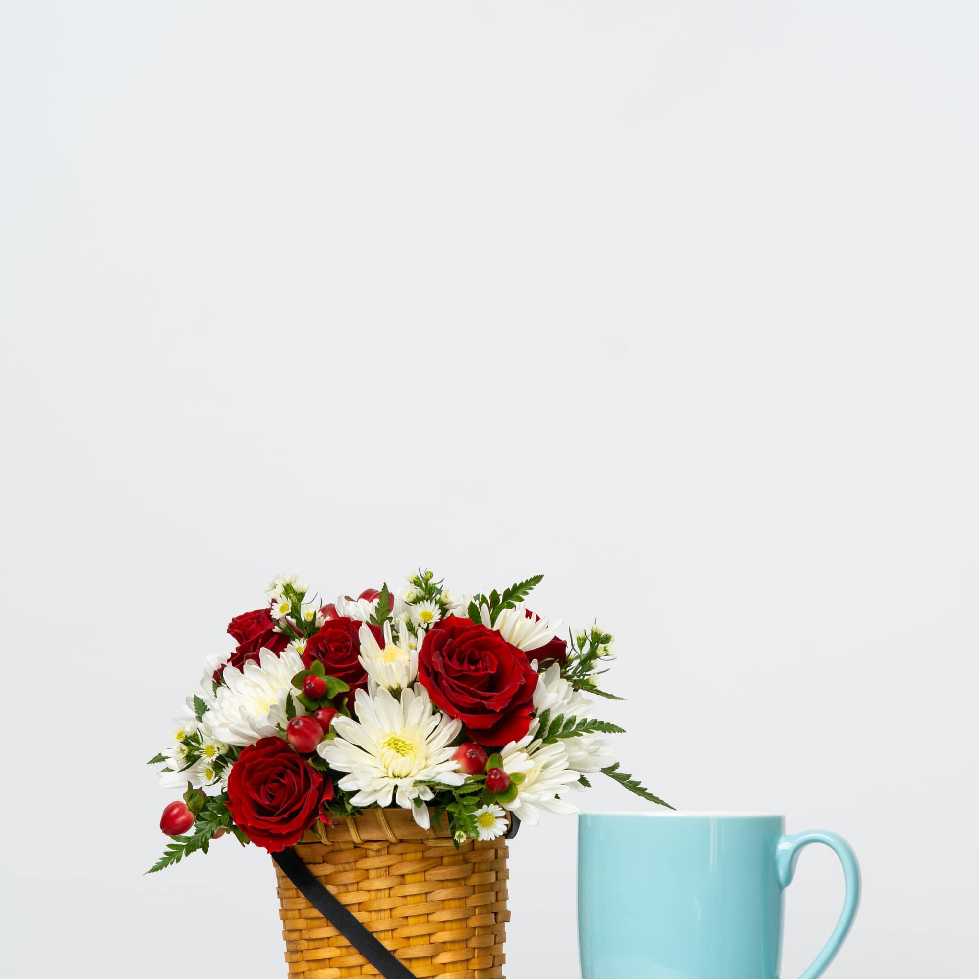 Red roses and white daisies in a woven basket beside a blue mug