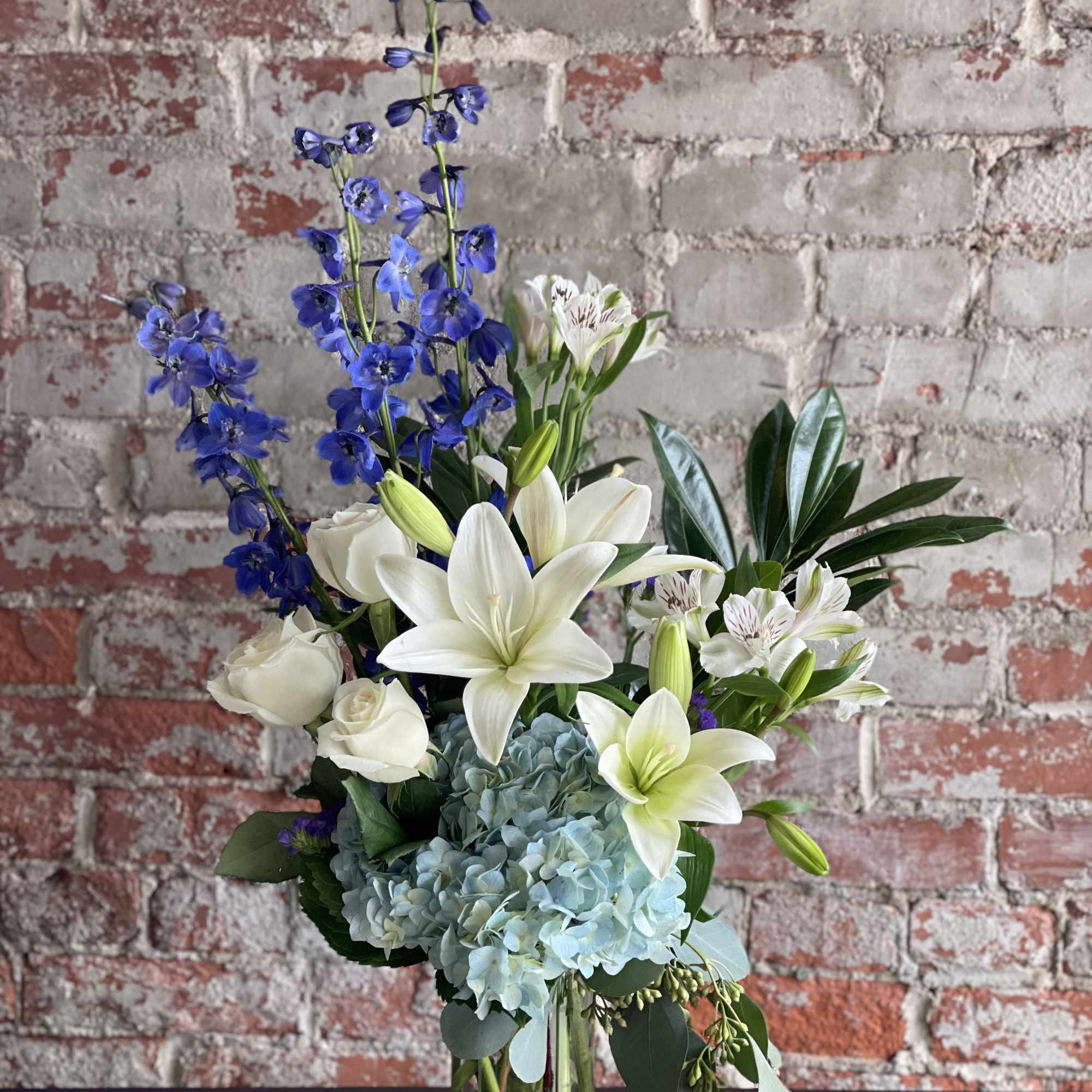 Tall bouquet of white lilies, blue flowers, and pale hydrangeas in a glass vase
