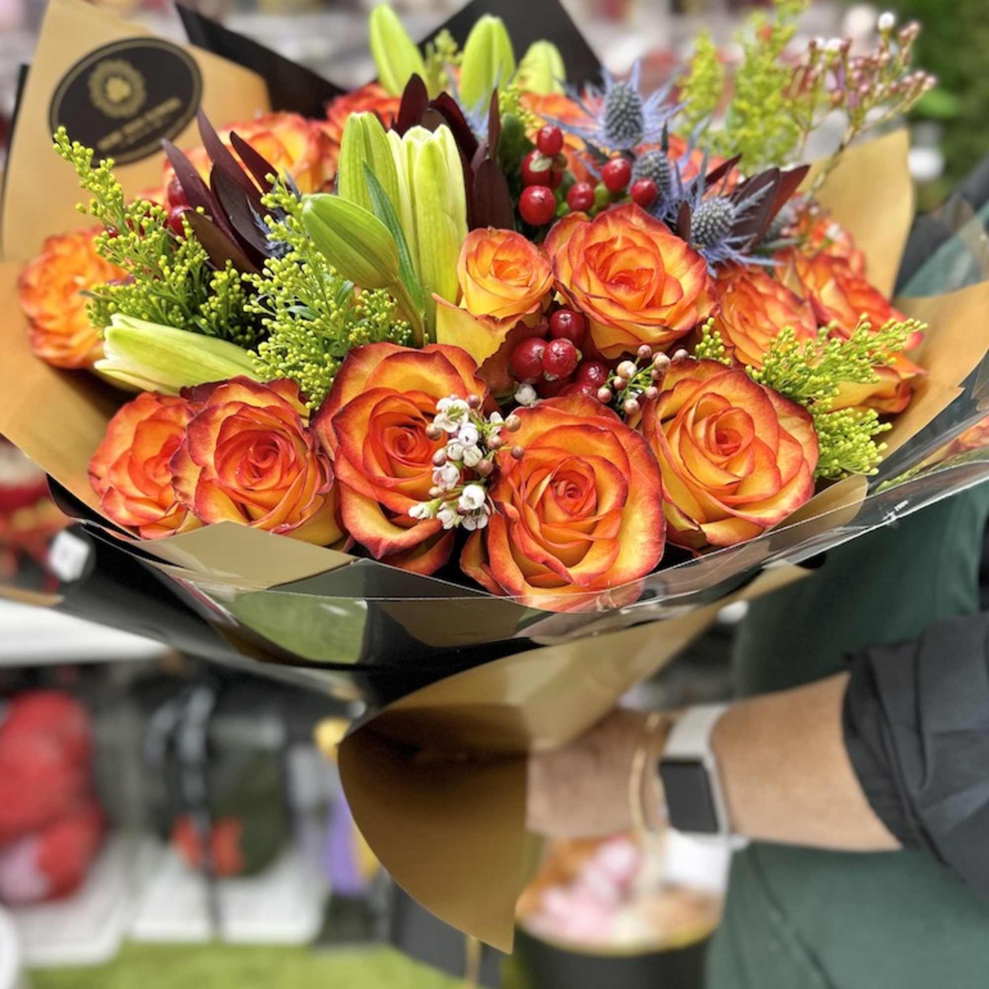 Hand-tied bouquet of orange-yellow roses with lilies and berries wrapped in black and gold paper