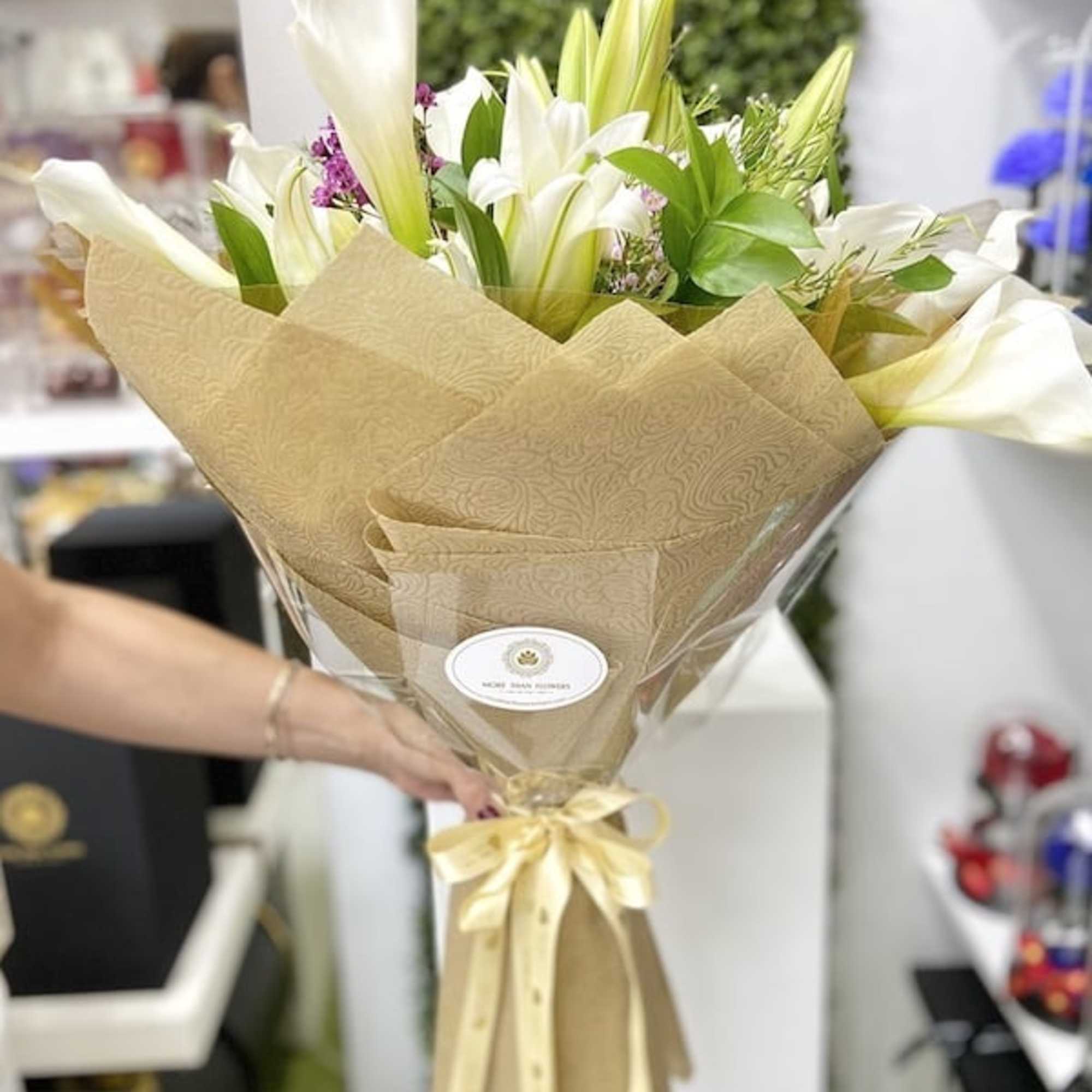 Wrapped bouquet of white lilies and calla lilies with touches of purple flowers in tan paper and a yellow bow