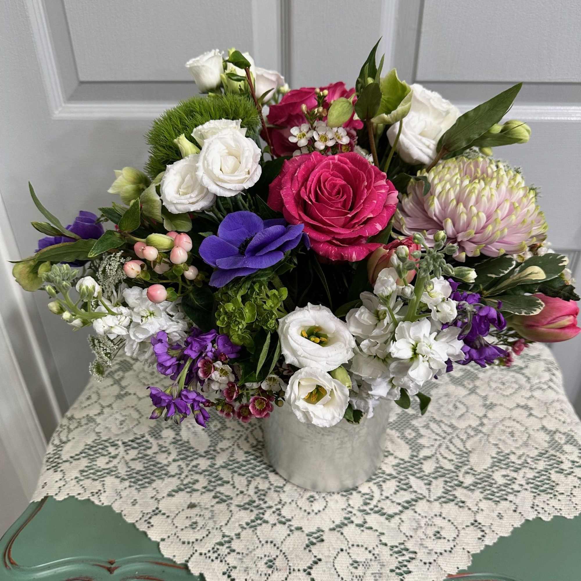 Mixed arrangement of pink roses, white lisianthus, and purple blooms in a metal vase on a lace-covered table