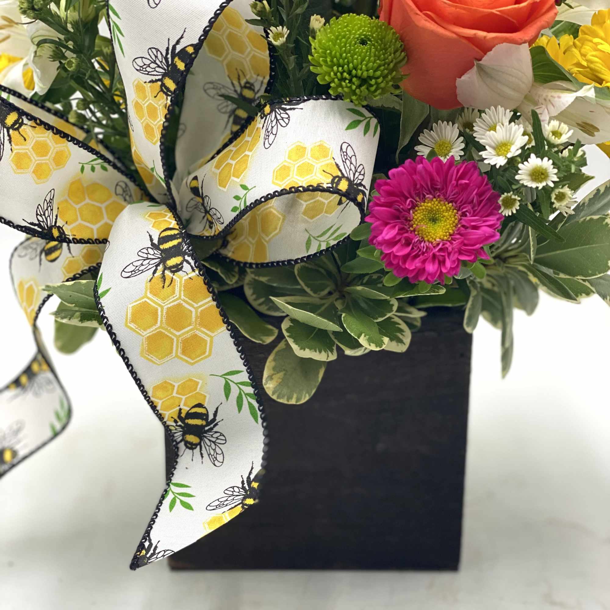 Flower arrangement in a black square container with a bee-patterned ribbon