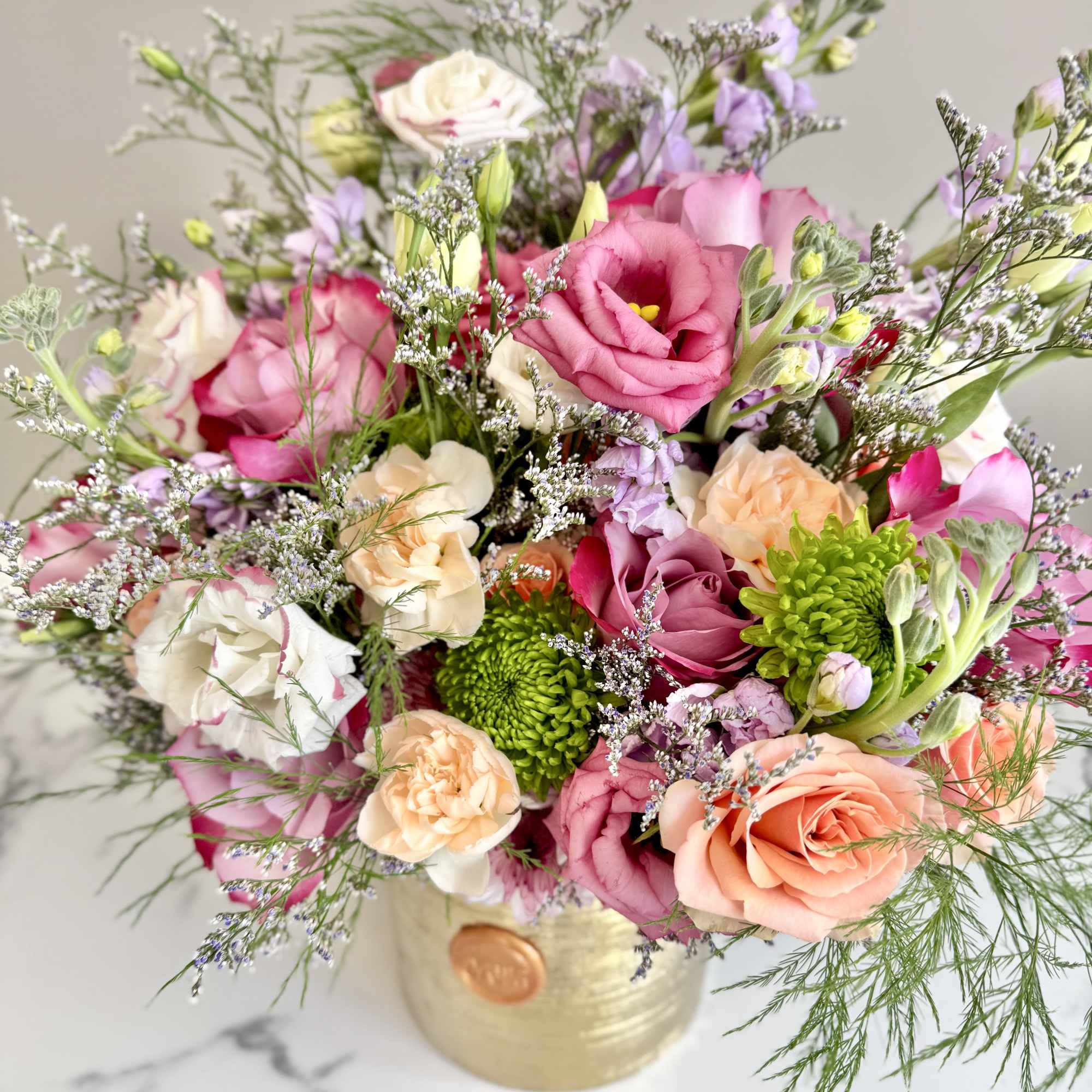 Arrangement of pink and peach roses, lisianthus, and green mums in a gold cylinder vase with airy greenery.