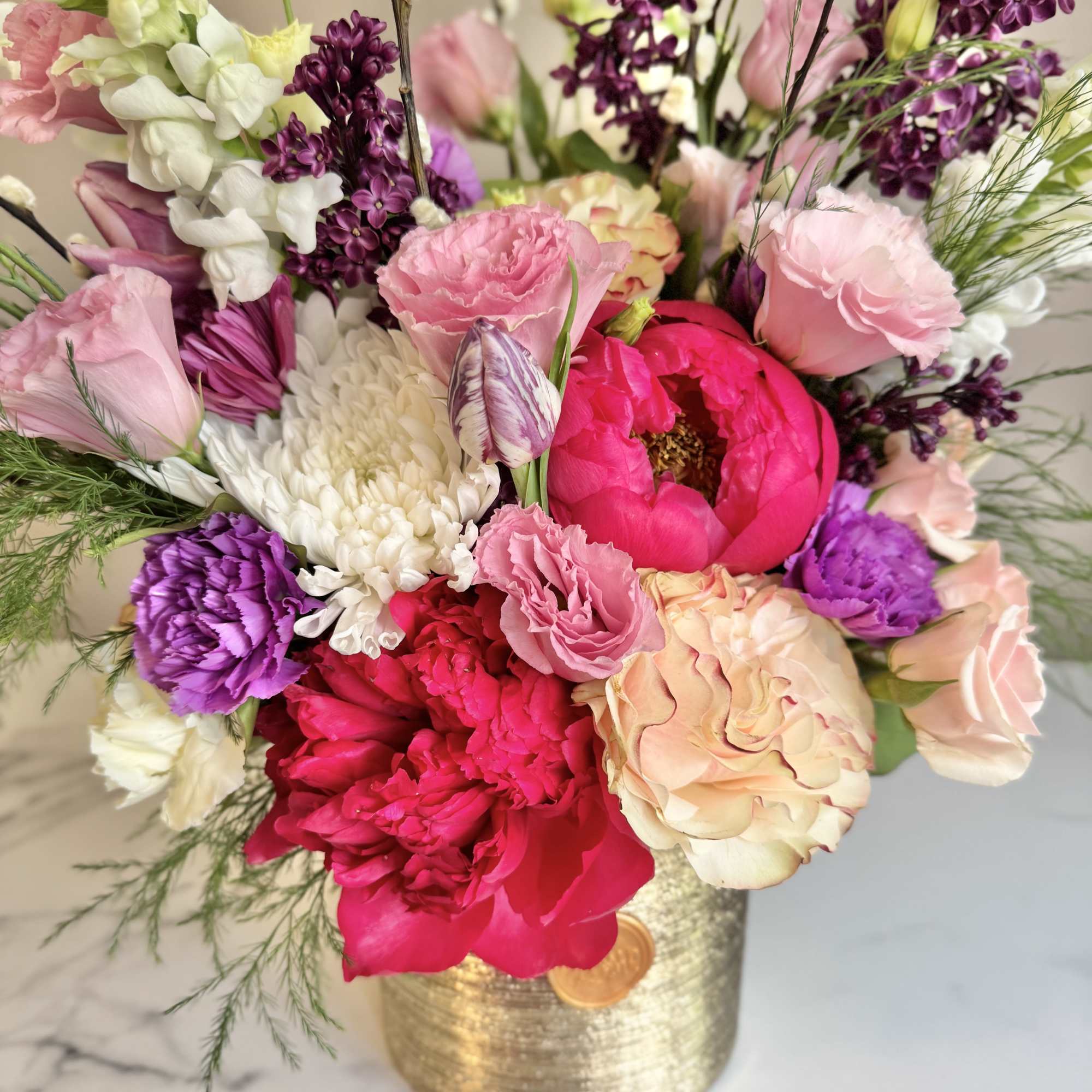 Lush arrangement of pink peonies, roses, purple blooms and white mums in a gold vase.