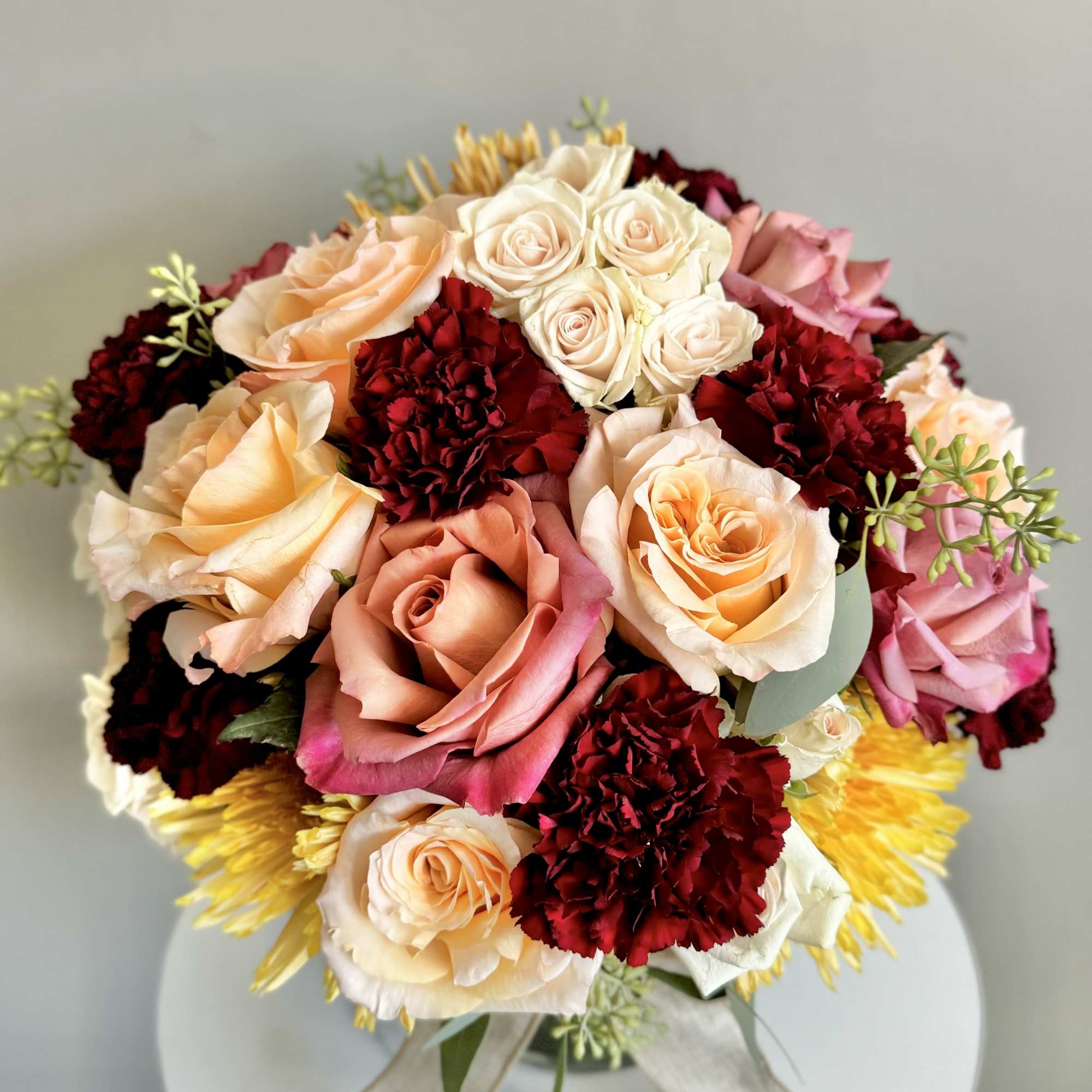 Round arrangement of peach roses, red carnations, and yellow mums in a vase with a ribbon bow.