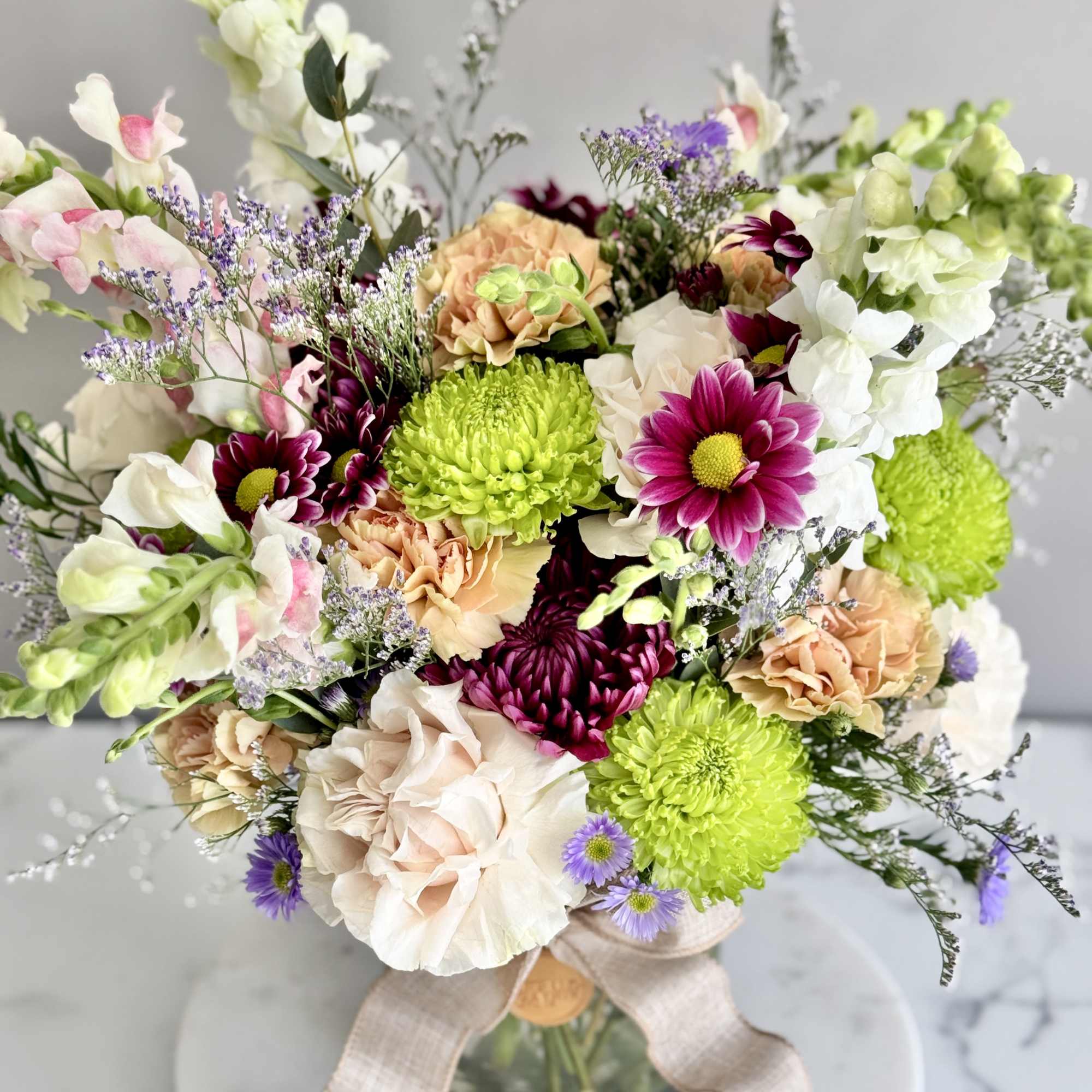 Mixed bouquet of green mums, peach blooms, and purple accents in a glass vase with a bow