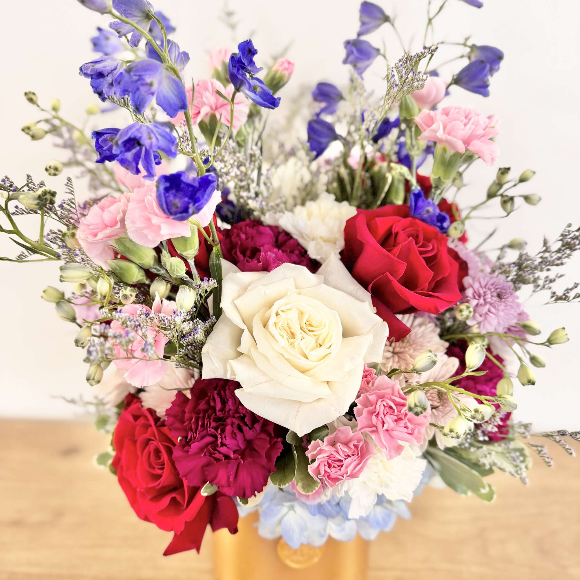 Mixed arrangement of red and white roses, pink carnations, and blue delphinium in a gold cylinder vase