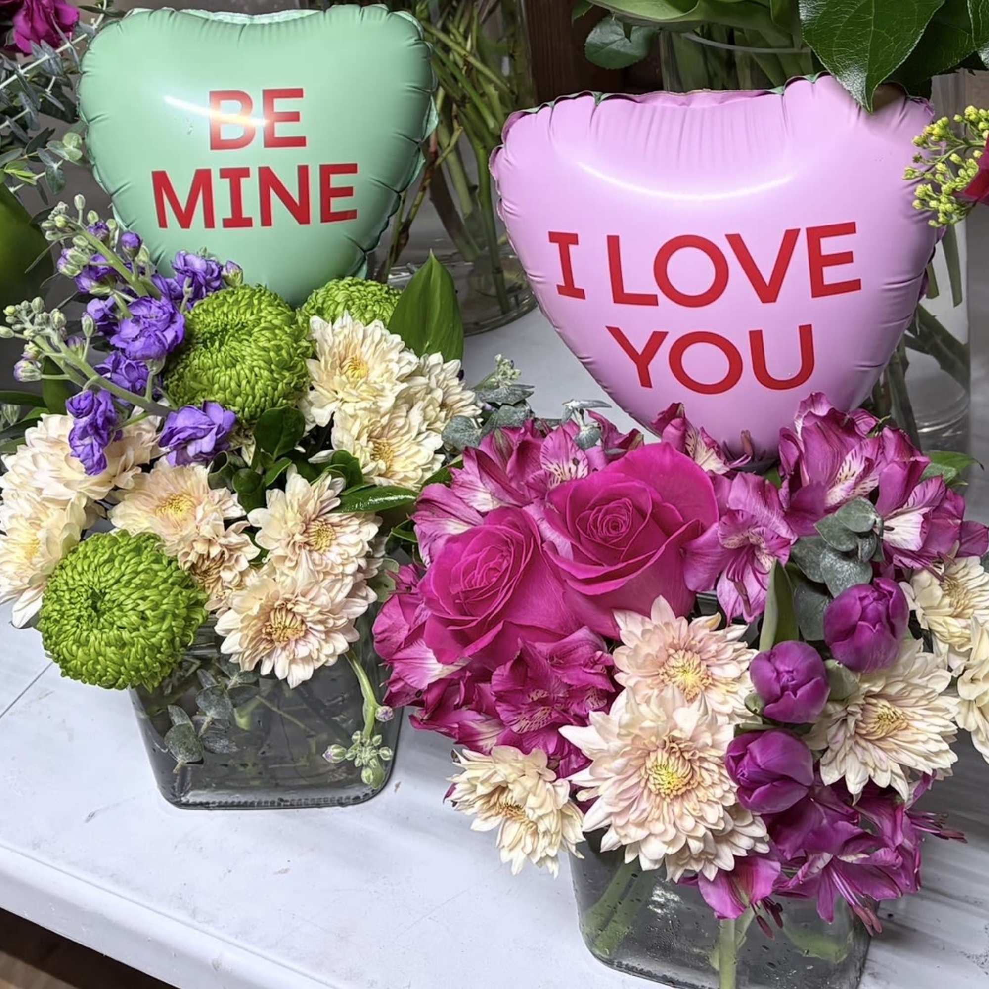 Two floral arrangements with heart balloons reading "BE MINE" and "I LOVE YOU"