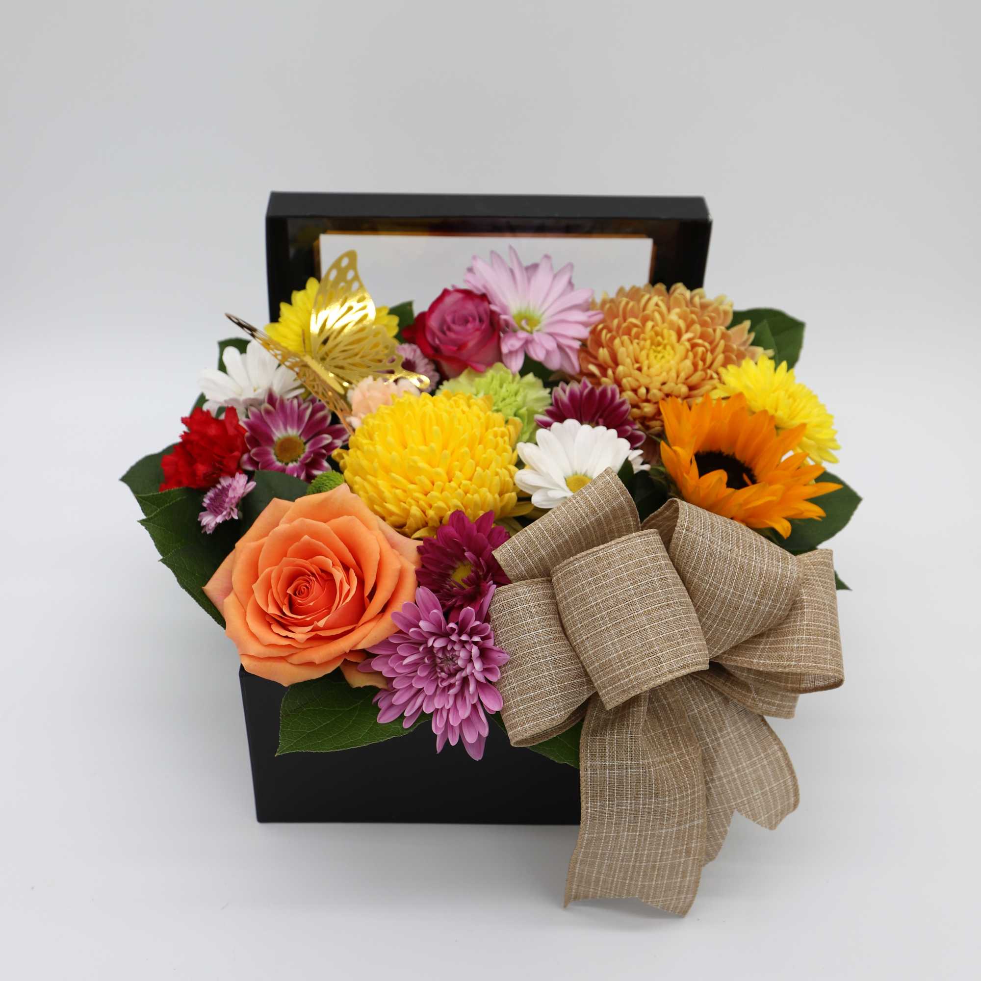 Mixed flowers arranged in a black box with a large tan ribbon bow