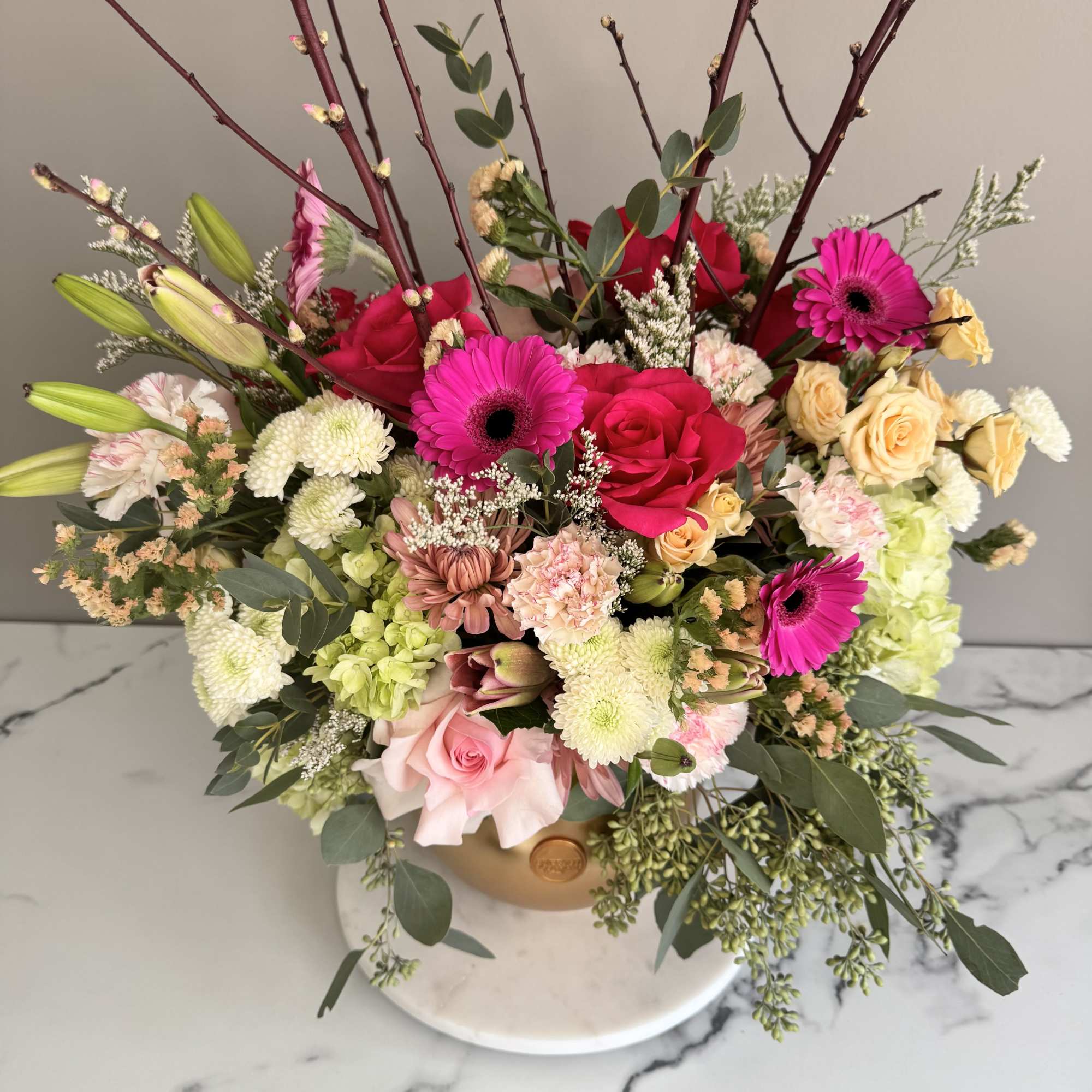 Tall mixed arrangement of pink gerberas, red and pink roses, lilies, and mums in a gold hat box.