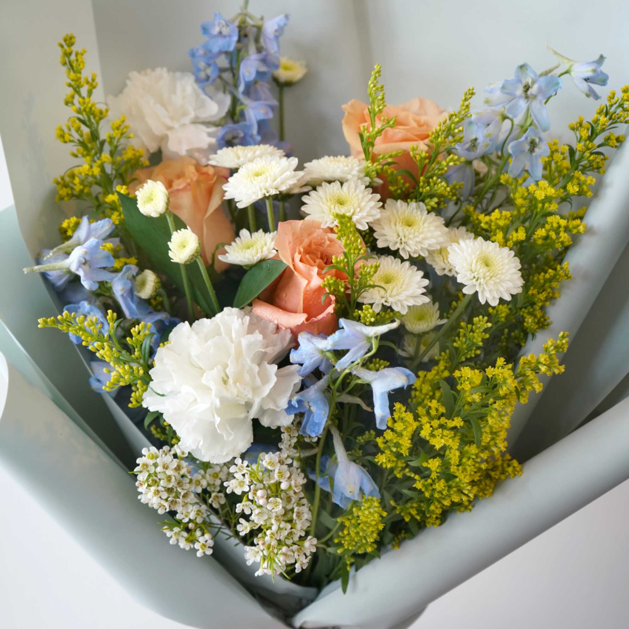 Wrapped bouquet of peach roses, white carnations and daisies with blue delphinium and yellow filler flowers.