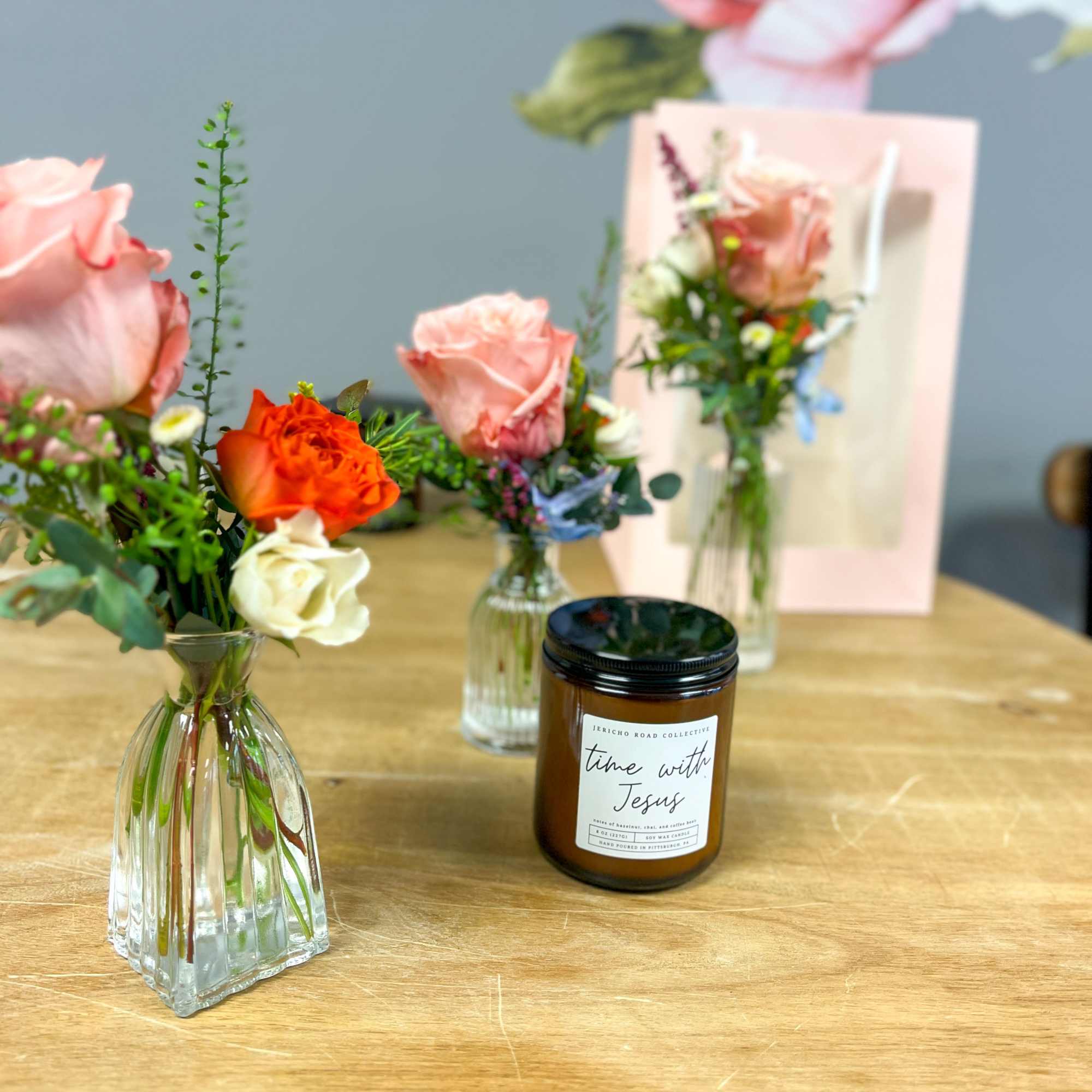 Small bud vases with pink and orange roses arranged around an amber glass candle on a wooden table