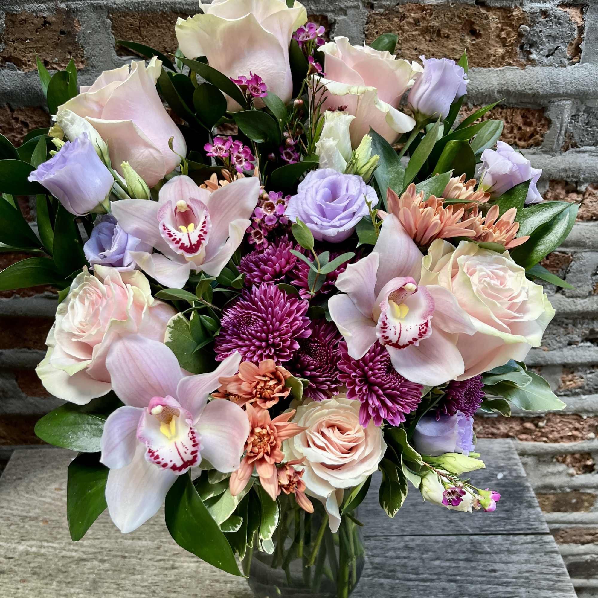 Vase arrangement of pink roses, lavender blooms, and orchids with magenta mums against a brick wall