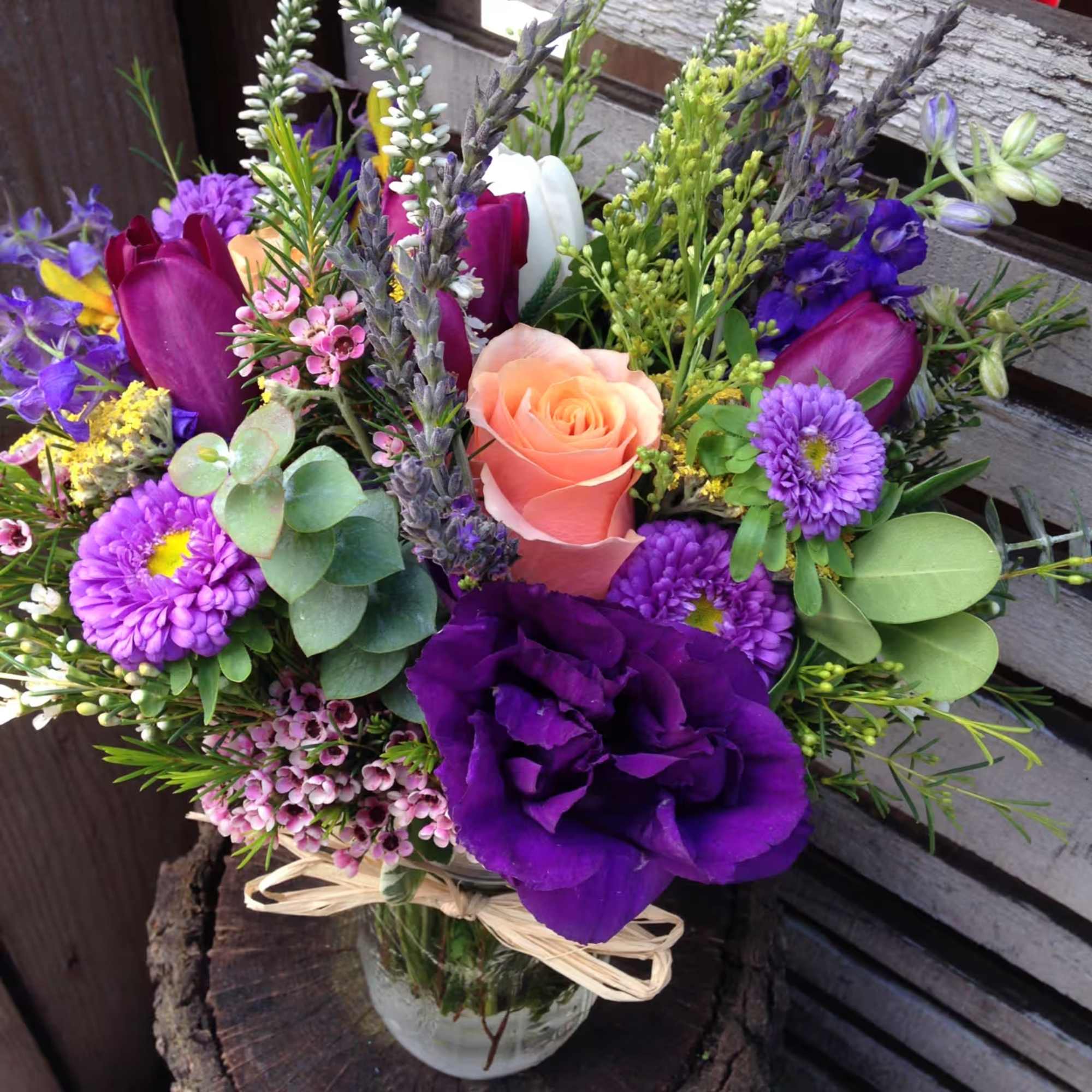 Colorful arrangement of peach roses and purple blooms in a clear glass jar tied with raffia