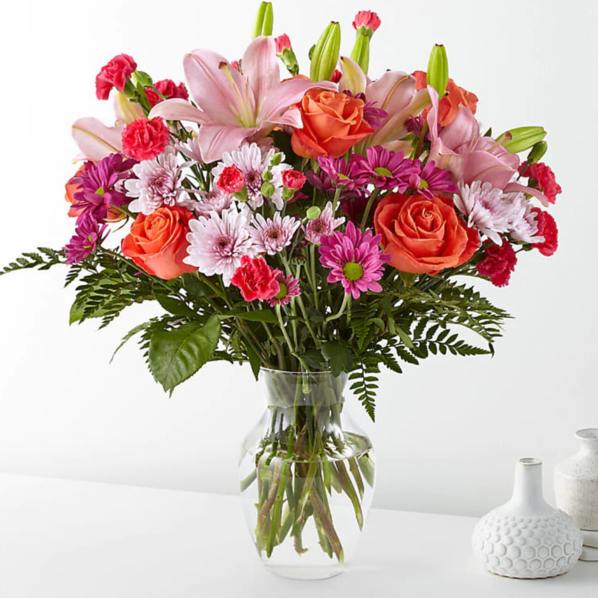 Mixed bouquet of pink lilies, orange roses, and daisies in a clear glass vase