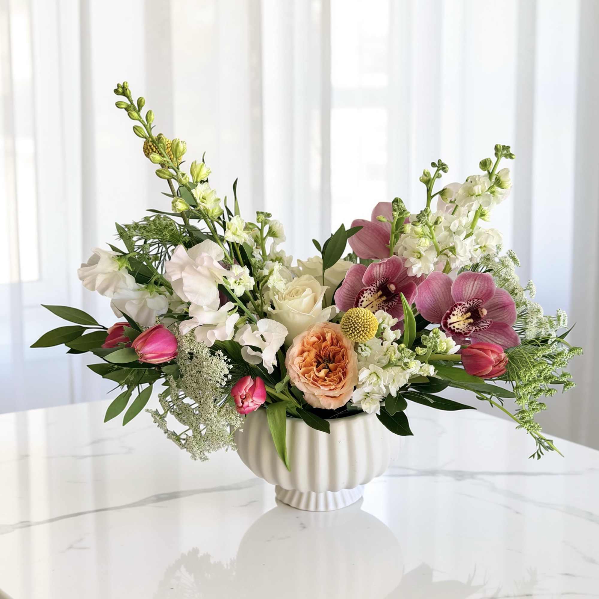 Low arrangement of pink orchids, tulips, and white blooms in a white ceramic bowl vase