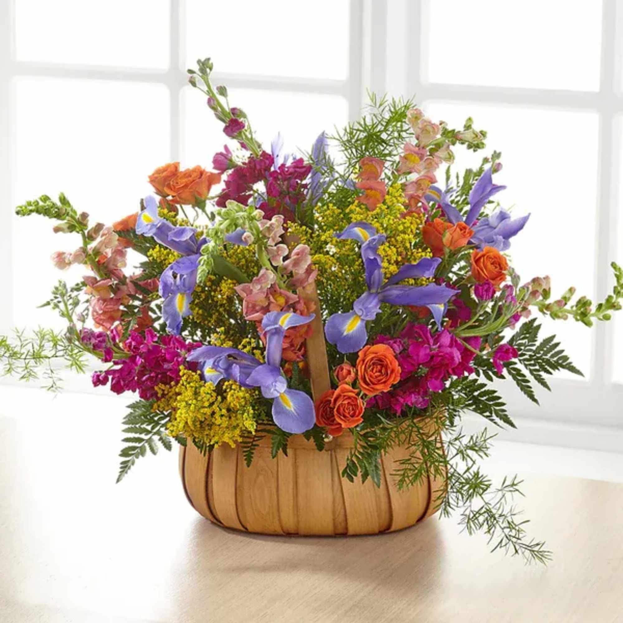 Bright mixed flower basket with purple irises, orange roses, and yellow blooms in a wooden handled basket.