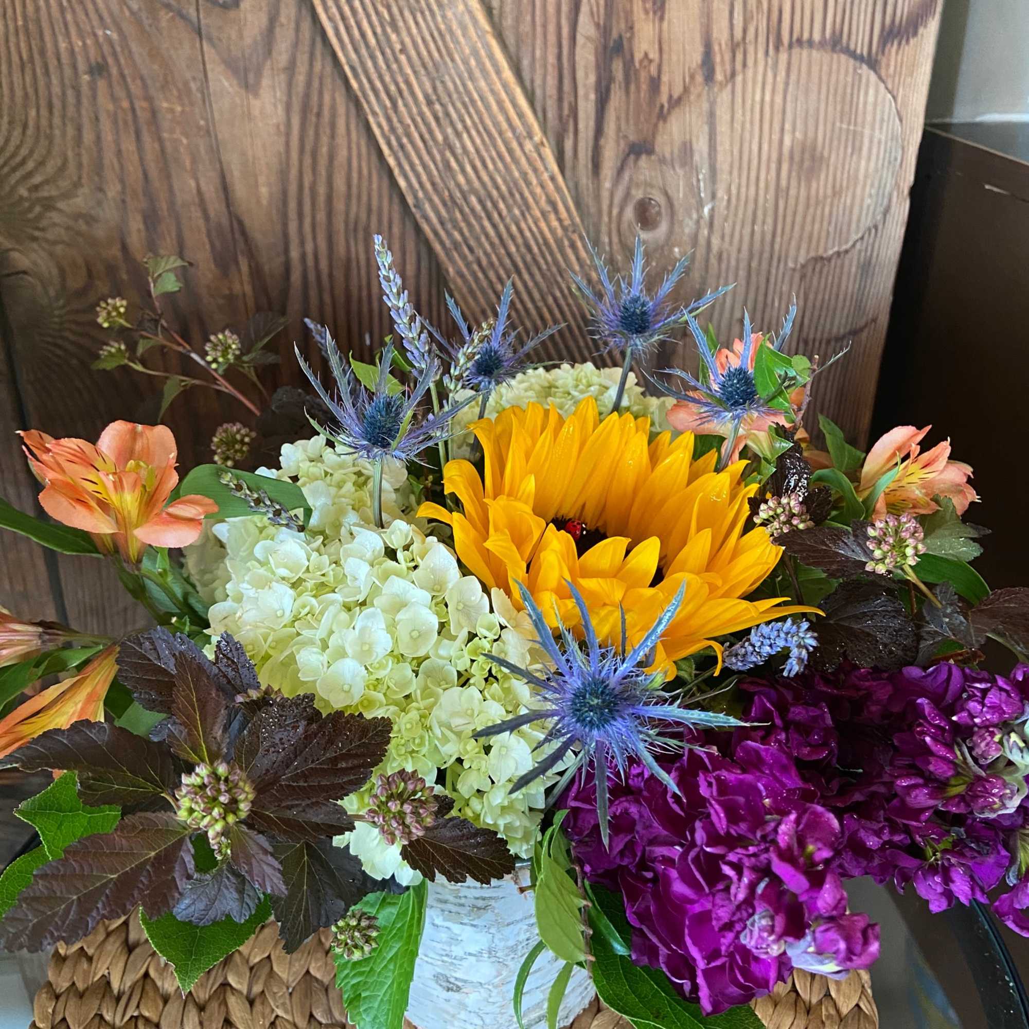 Rustic arrangement with yellow sunflower, white hydrangea, and purple blooms in a birch-wrapped vase