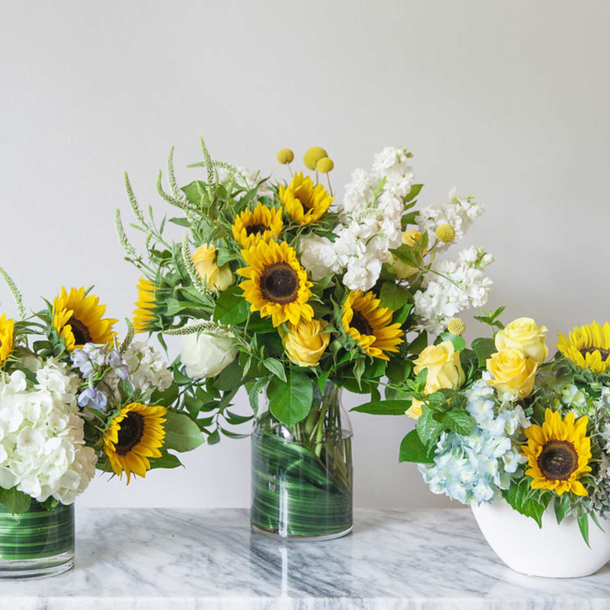 Three floral arrangements with sunflowers and white blooms in glass and white containers