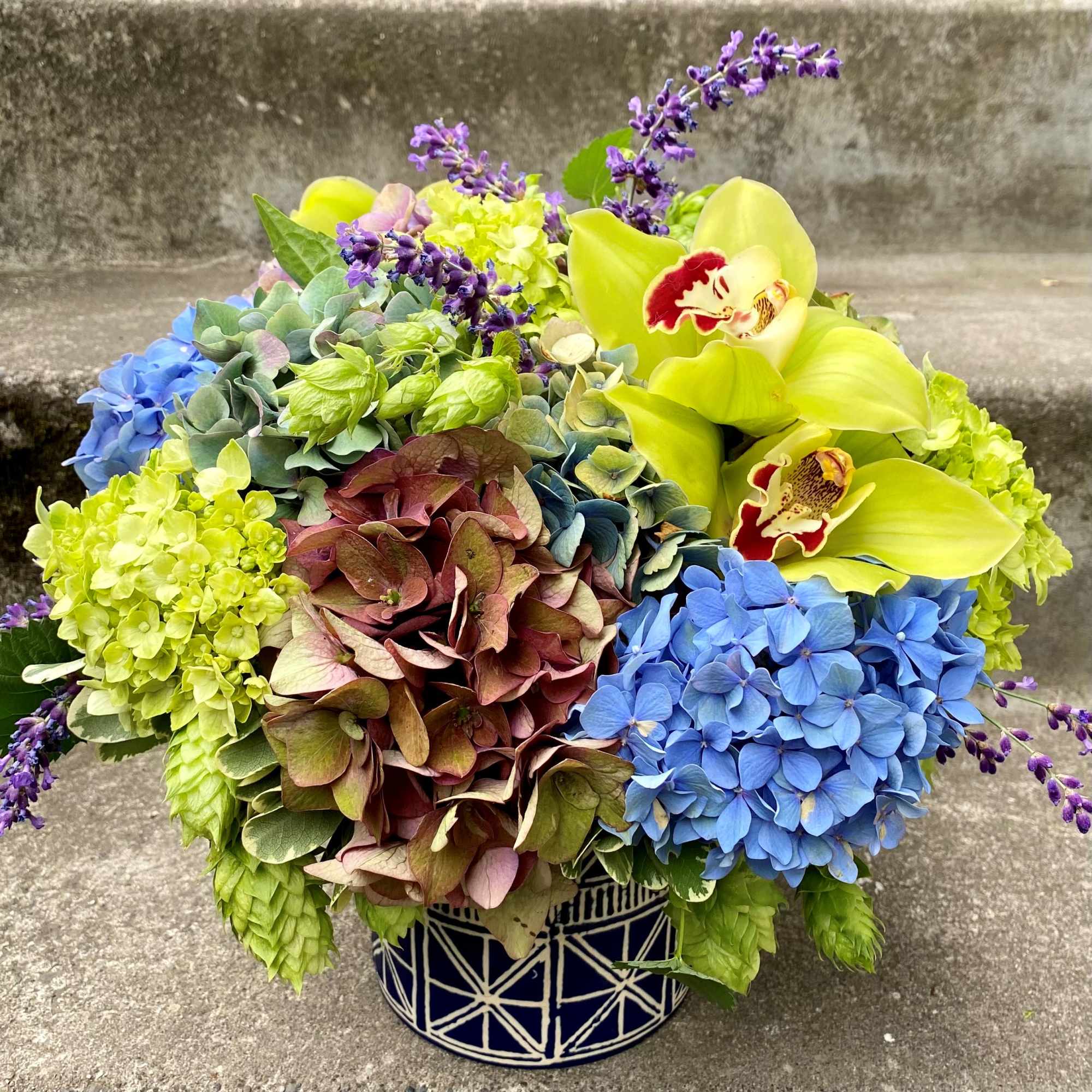 Colorful bouquet with hydrangeas, orchids, and purple accents in a patterned vase