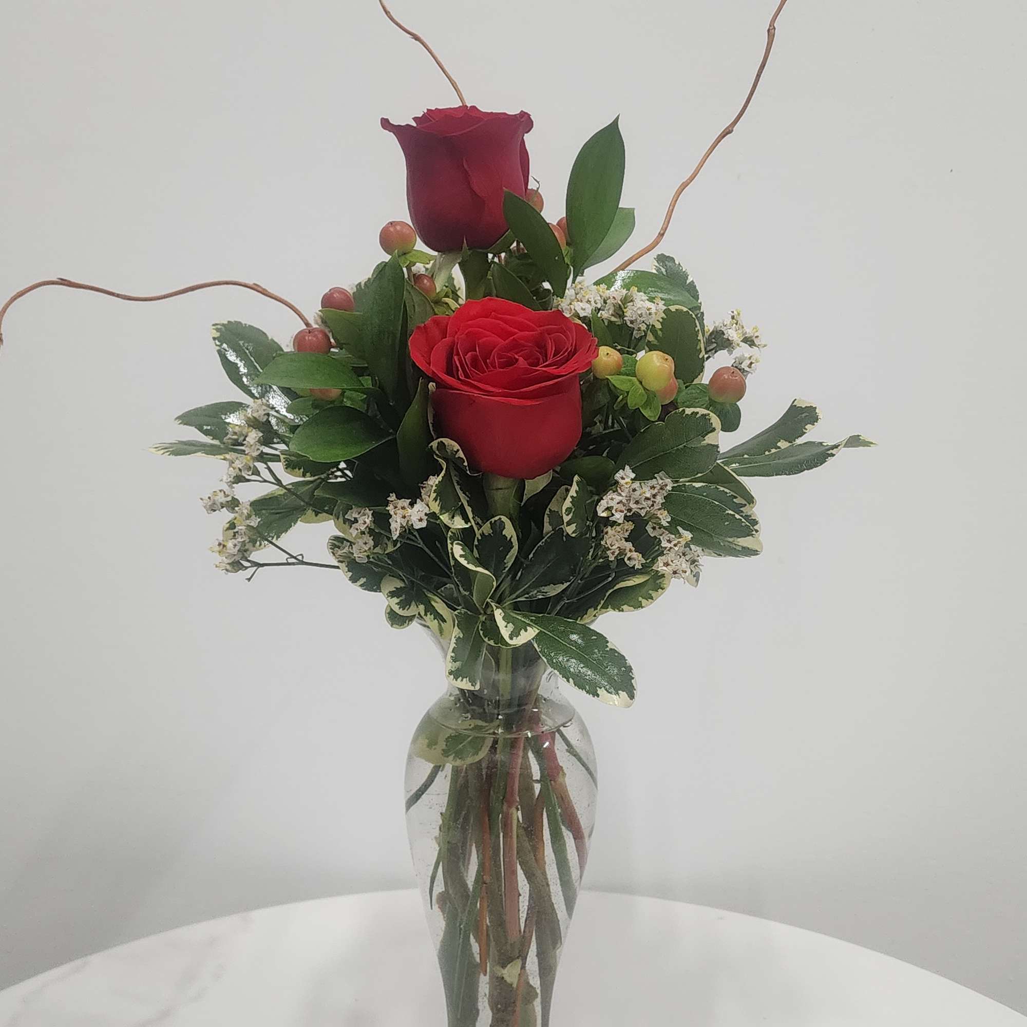Two red roses in a glass vase with greenery and small white filler flowers