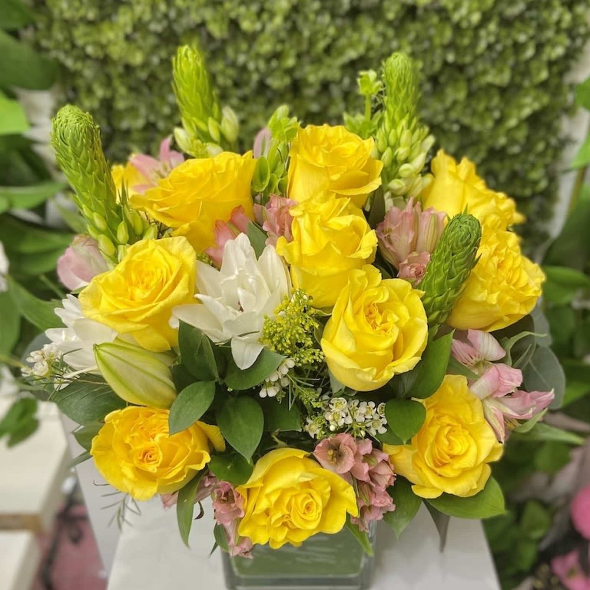 Low arrangement of yellow roses, white lilies and pink flowers in a clear square glass vase