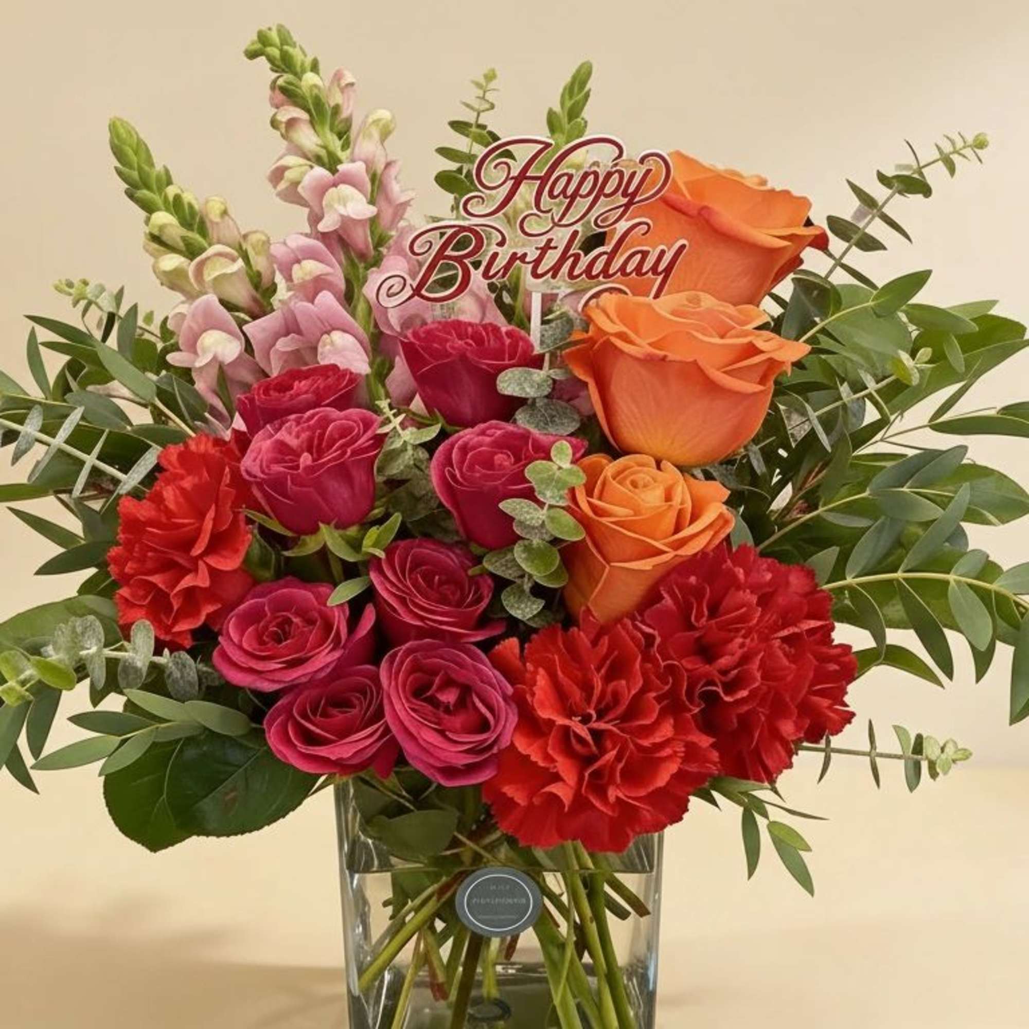 Bright orange roses, pink roses, and red carnations in a glass vase with a Happy Birthday sign.