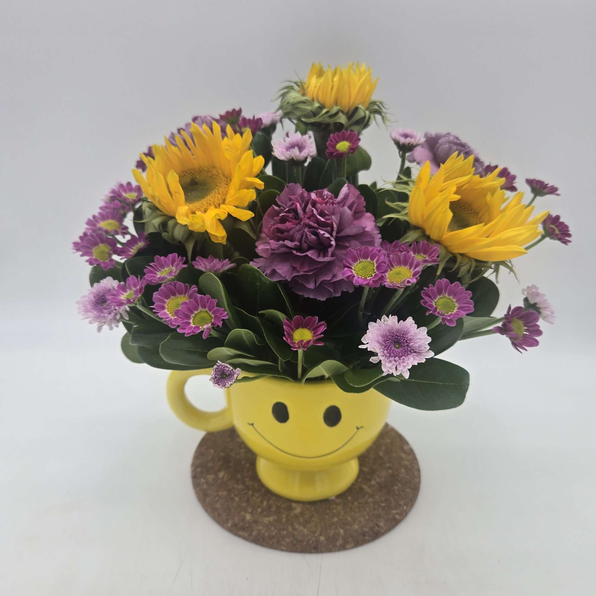 Yellow sunflower and purple daisy bouquet in a smiley-face mug